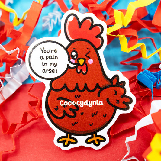 A red chicken cartoon sticker with a cheeky wink and speech bubble saying “You’re a pain in my arse!” by Innabox. The fun design reads “Cock-cydynia” and sits among colourful red, yellow, and blue paper confetti. This glossy vinyl sticker is waterproof, about 5 cm tall, and perfect for adding humour to laptops, water bottles, or journals. Disability-inspired and made to spread joy.
