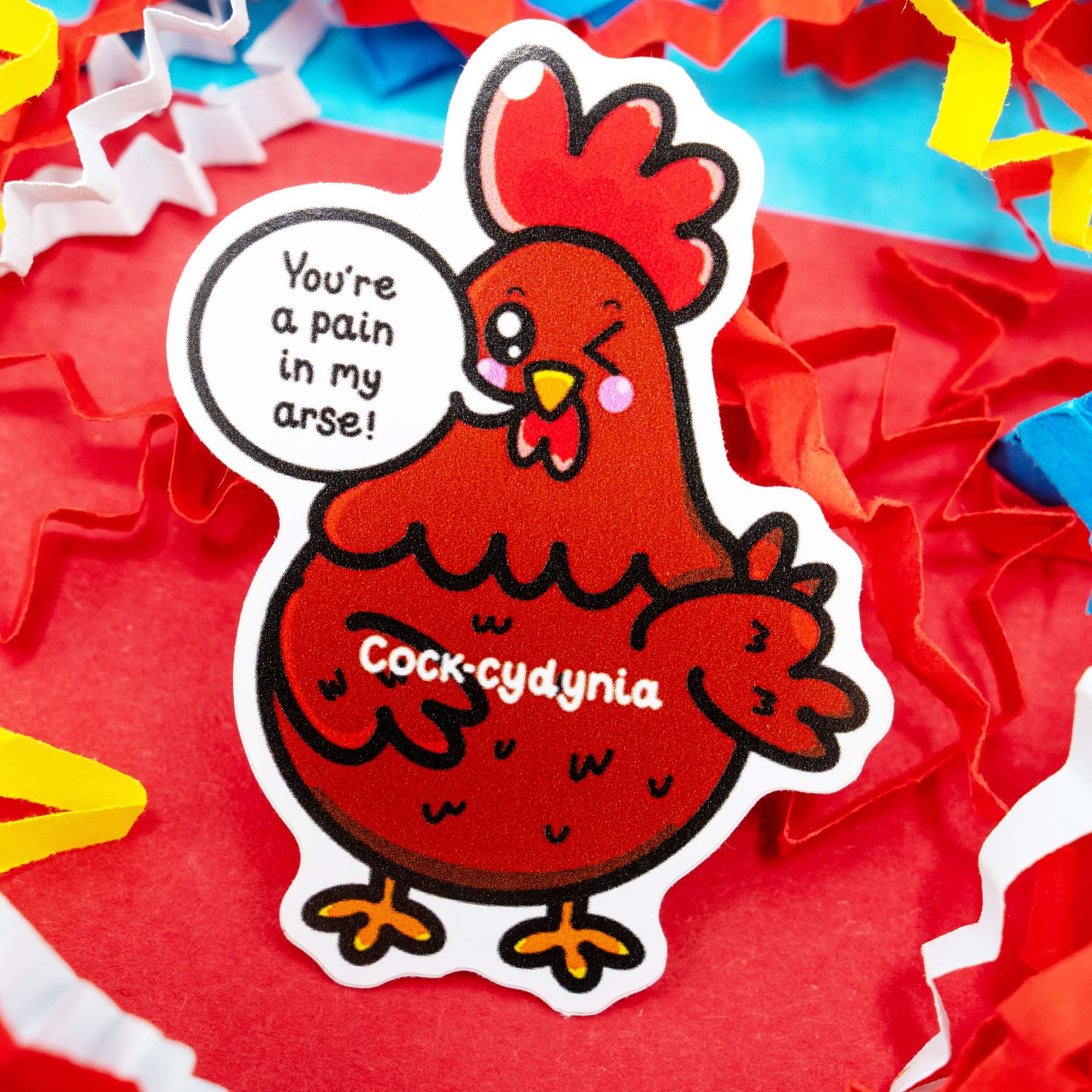 Funny red chicken vinyl sticker with the phrase “You’re a pain in my arse!” by Innabox. Features a winking chicken with “Cock-cydynia” text and bright crinkle paper background. Gloss vinyl finish, waterproof, and around 5 cm tall. Ideal for laptops, water bottles, or journals. Disability-inspired design made to bring laughter and kindness to your day.