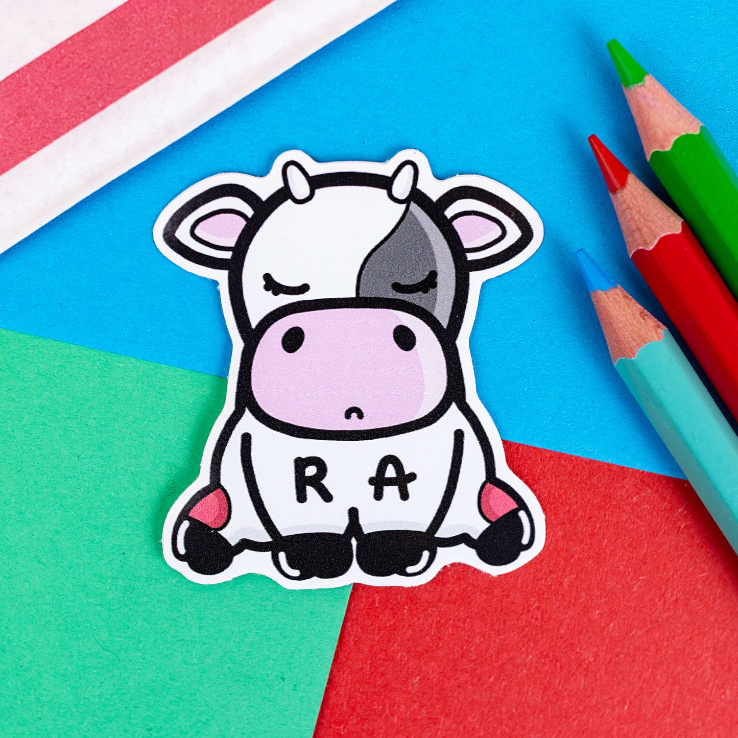 The Moomatoid Arthritis Cow Sticker - Rheumatoid Arthritis on a red, blue and green background with colouring pencils and red stripe candy bag. The sad cow shape sticker has red marks on its lower back legs and the initials R and A across its middle. The hand drawn design is raising awareness for Rheumatoid Arthritis.