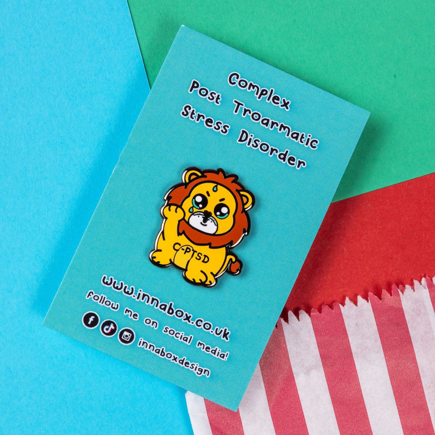 The Complex Post Troarmatic Stress Disorder Lion Enamel Pin - C-PTSD - Complex Post Traumatic Stress Disorder on blue backing card with black text. A yellow crying smiling and sweating male lion with its paw raised in the middle with 'C-PTSD' across its chest. The enamel pin design is raising awareness for complex post traumatic stress disorder.