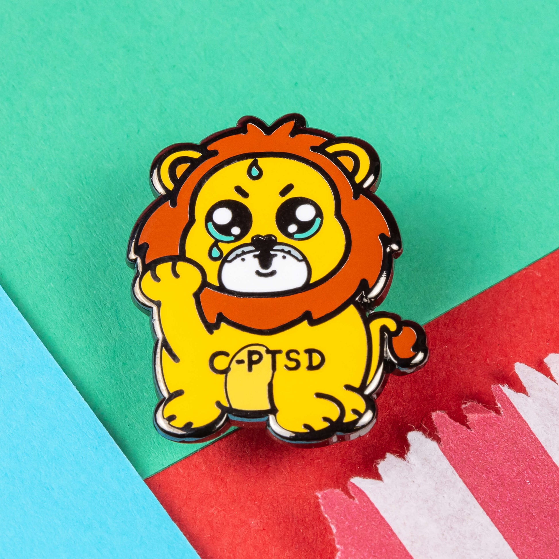 The Complex Post Troarmatic Stress Disorder Lion Enamel Pin - C-PTSD - Complex Post Traumatic Stress Disorder on a red, blue and green card background. A yellow crying smiling and sweating male lion with its paw raised in the middle with 'C-PTSD' across its chest. The enamel pin design is raising awareness for complex post traumatic stress disorder.