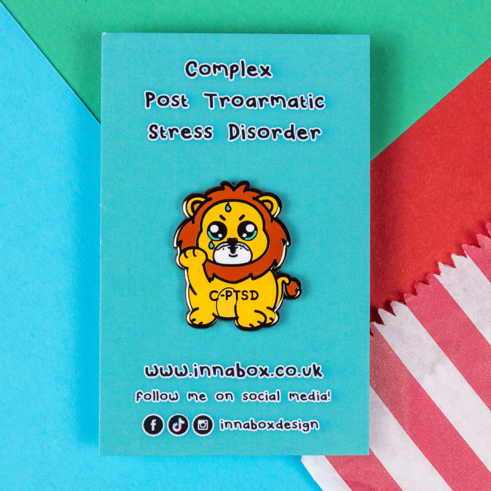 The Complex Post Troarmatic Stress Disorder Lion Enamel Pin - C-PTSD - Complex Post Traumatic Stress Disorder on blue backing card with black text. A yellow crying smiling and sweating male lion with its paw raised in the middle with 'C-PTSD' across its chest. The enamel pin design is raising awareness for complex post traumatic stress disorder.