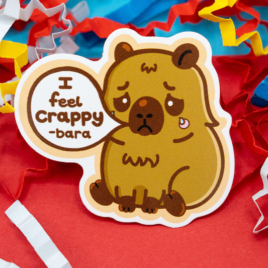 A cute brown capybara cartoon sticker with a sad face and a tear, saying “I feel crappy-bara” inside a speech bubble. Surrounded by red, yellow and blue paper confetti, the design by Innabox raises awareness for mental health and chronic illness through humour and kindness. Glossy vinyl, waterproof and around 5 cm tall, this disability-inspired sticker adds a gentle smile to your day.