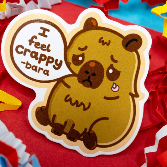 Adorable capybara vinyl sticker by Innabox with the phrase “I feel crappy-bara.” Features a sad brown capybara cartoon with a tear and colourful paper background. Gloss vinyl, waterproof, around 5 cm tall and custom cut. A thoughtful mental health sticker designed to comfort and uplift those with chronic illness or hidden disabilities.