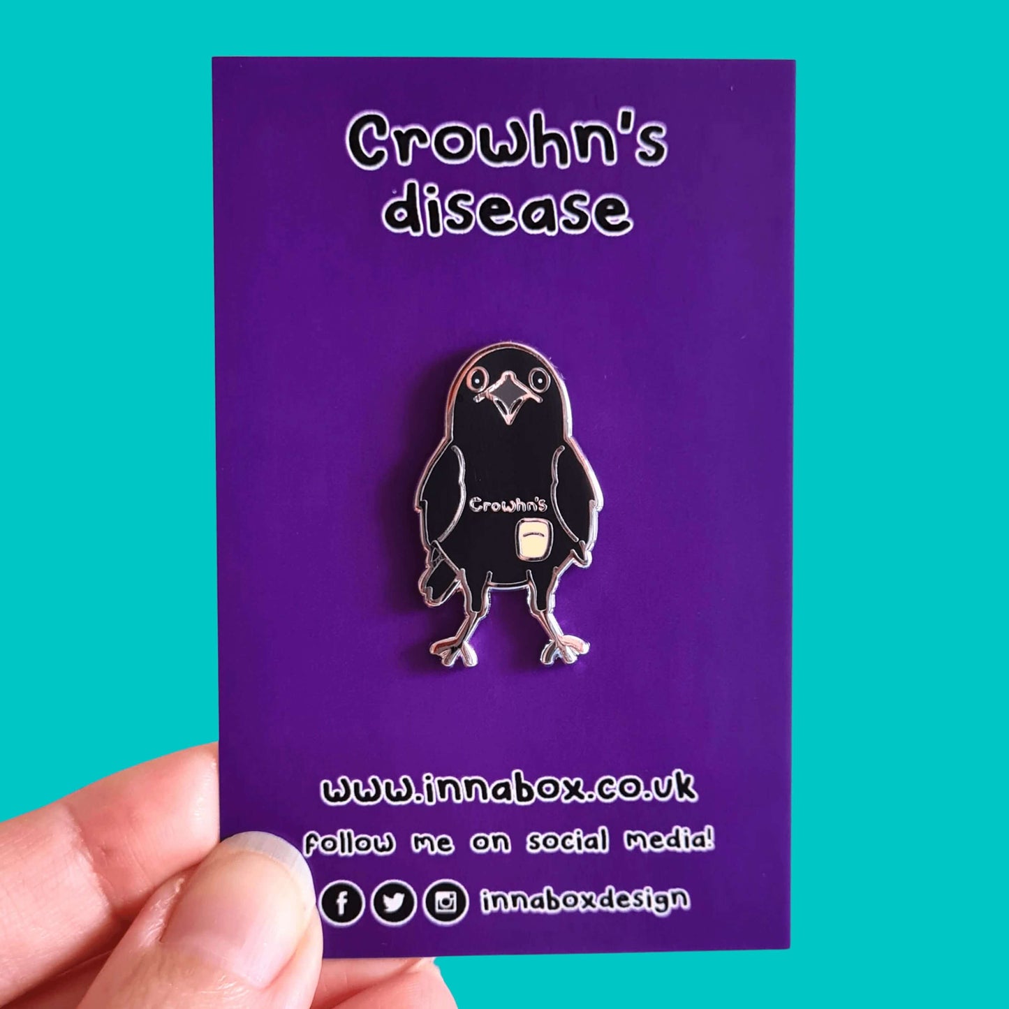 The Crowhn's Disease Enamel Pin - Crohn's Disease on purple backing card with top text reading 'Crowhn's disease' and bottom text of innabox social media handles being held over a blue background. The black crow shape pin has a white stoma bag fitted and the text reading 'chrowhn's' across its chest. The design is raising awareness for crohn's disease.