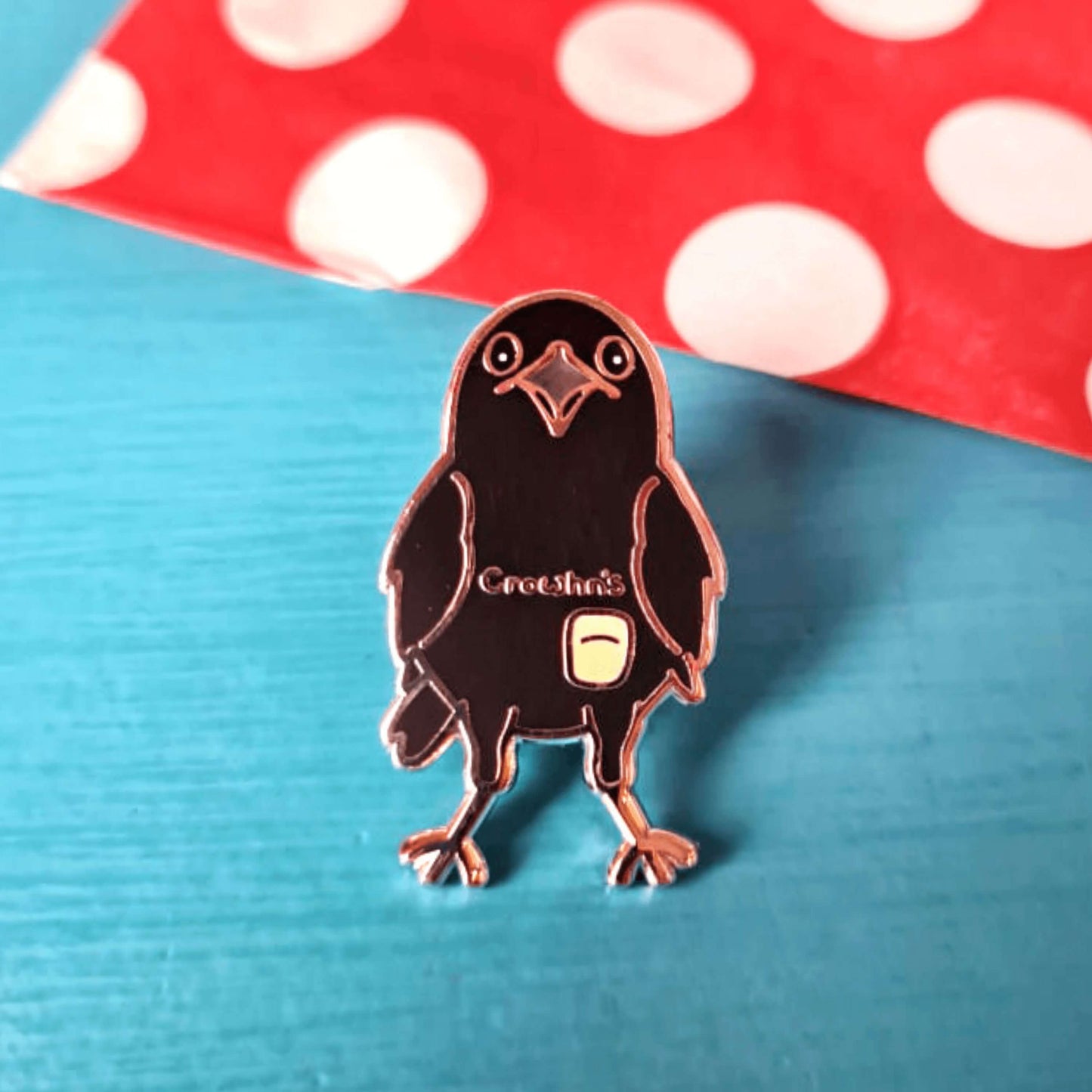 The Crowhn's Disease Enamel Pin - Crohn's Disease on a red and blue background. The black crow shape pin has a white stoma bag fitted and the text reading 'chrowhn's' across its chest. The design is raising awareness for crohn's disease.