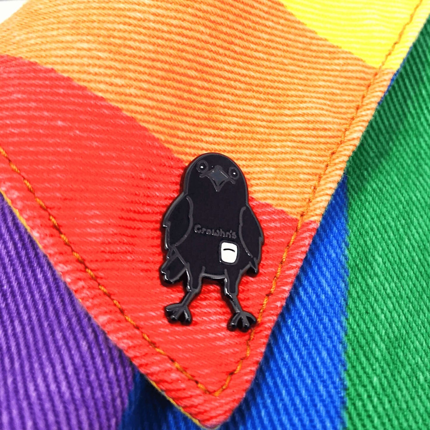 The Crowhn's Disease Enamel Pin - Crohn's Disease on a rainbow denim jacket collar. The black crow shape pin has a white stoma bag fitted and the text reading 'chrowhn's' across its chest. The design is raising awareness for crohn's disease.
