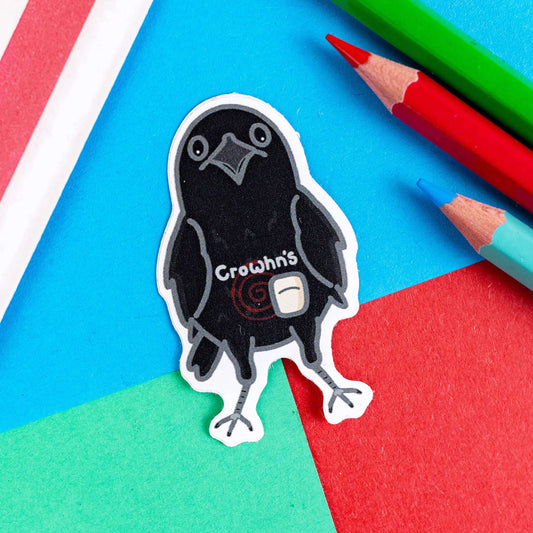 The Crowhn's Sticker - Crohn's Disease on a red, green and blue background with colouring pencils and a red stripe candy bag. The black crow shaped sticker has a stoma bag fitted with a red swirl and white text reading 'crowhn's' across its belly. The design is raising awareness for crohn's disease.