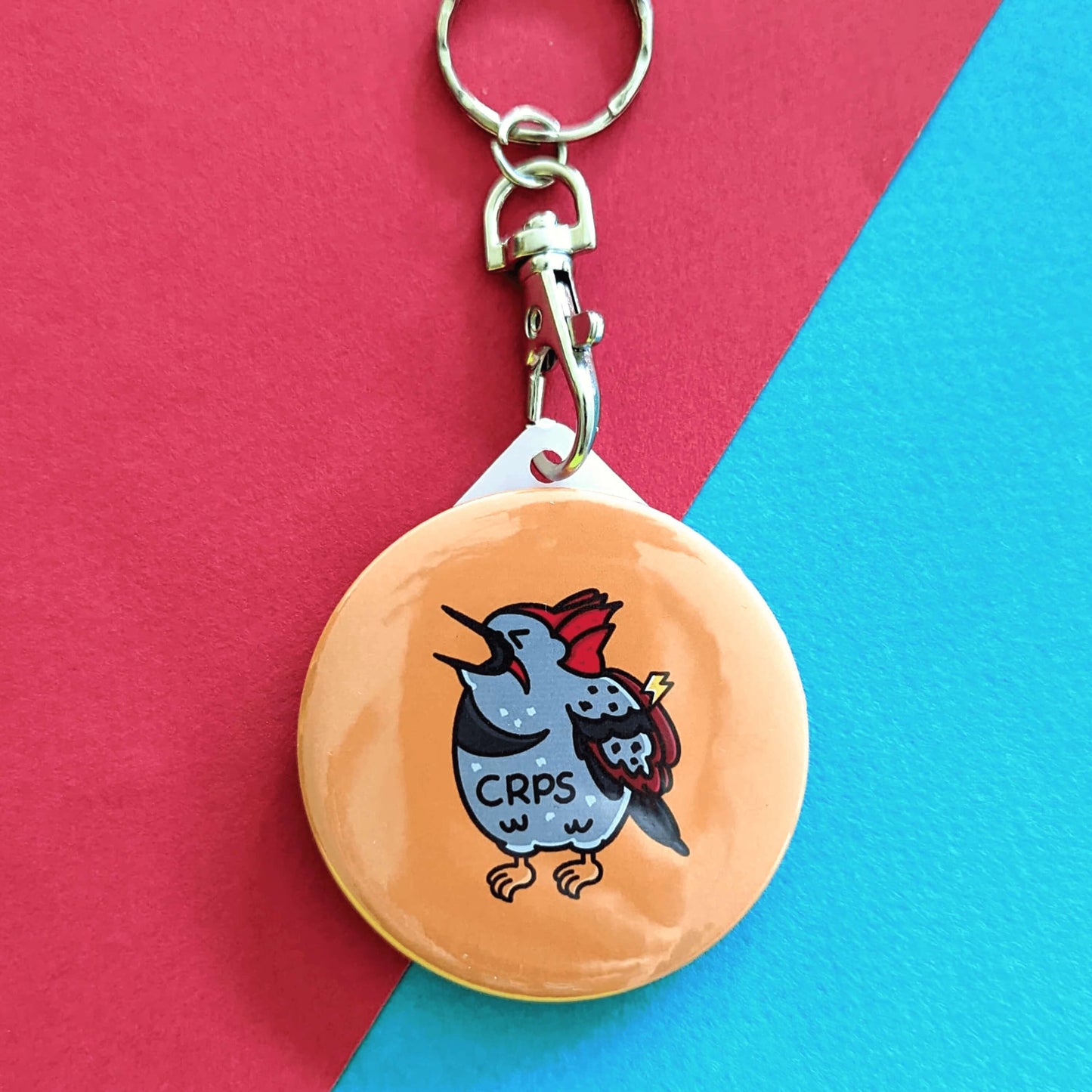 CRPS Keyring - Complex Regional Pain Syndrome on a red and blue background. The silver clip yellow plastic circular keychain features a screaming upset woodpecker bird with a lightning bolt on its wing and 'CRPS' written across its belly in black. The design is raising awareness for complex regional pain syndrome.
