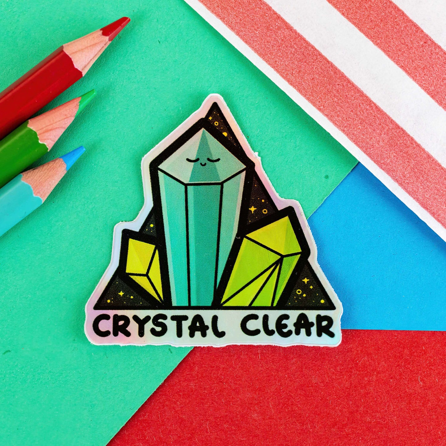 The Crystal Clear Holographic Sticker on a red, green and blue background with colouring pencils and a red stripe candy bag. The triangle shaped sticker features three green crystal towers on a black sparkly background with bottom text reading crystal clear, the centre crystal is smiling. Inspired by witchy spiritual healing with crystals.