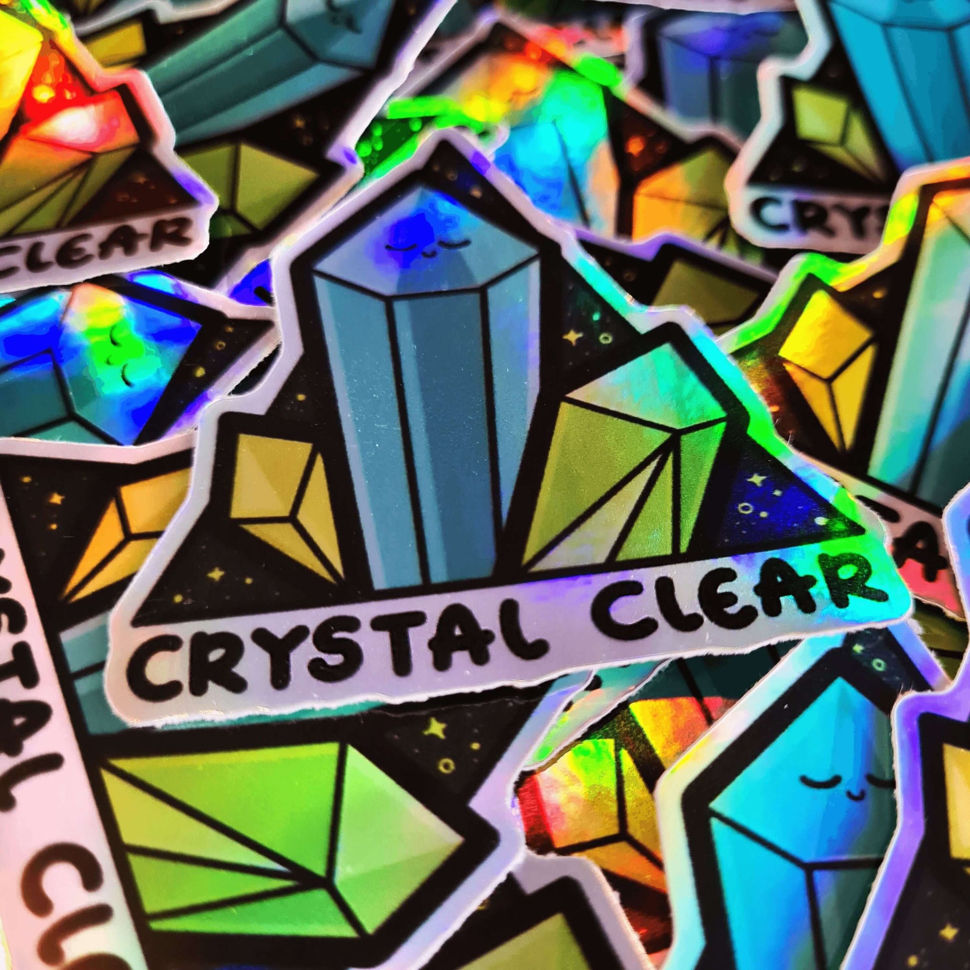 Multiple Crystal Clear Holographic Stickers in a pile. The triangle shaped sticker features three green crystal towers on a black sparkly background with bottom text reading crystal clear, the centre crystal is smiling. Inspired by witchy spiritual healing with crystals.