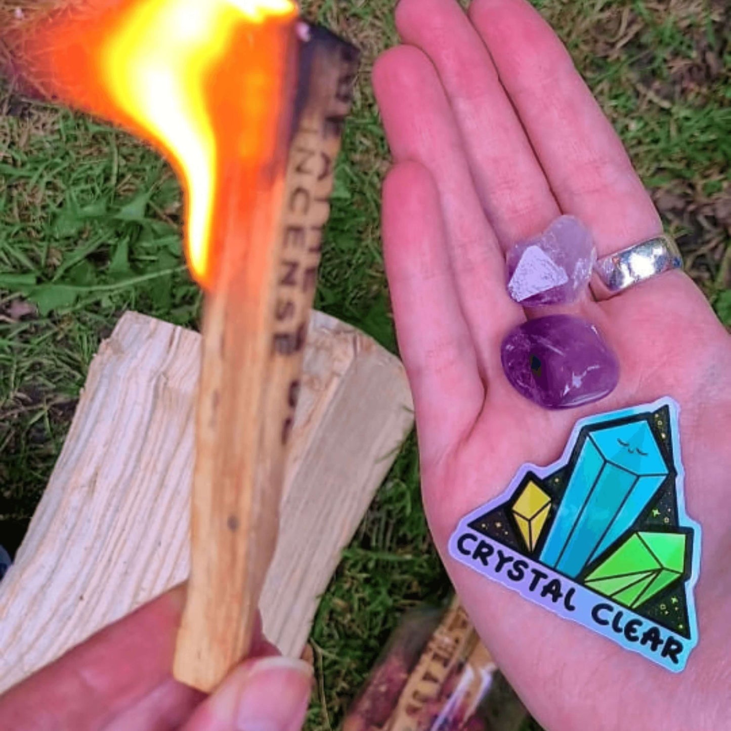 The Crystal Clear Holographic Sticker being held outside with a burnt incense stick and amethyst crystals. The triangle shaped sticker features three green crystal towers on a black sparkly background with bottom text reading crystal clear, the centre crystal is smiling. Inspired by witchy spiritual healing with crystals.