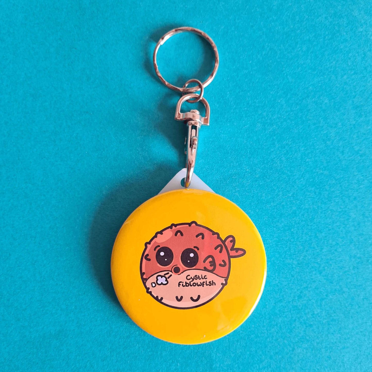 The Cystic Fiblowfish Keyring - Cystic Fibrosis on a blue background. The silver clip plastic yellow circle keychain with a puffer fish blowfish wheezing with 'cystic fiblowfish' written in black on its belly. The design is raising awareness for cystic fibrosis.
