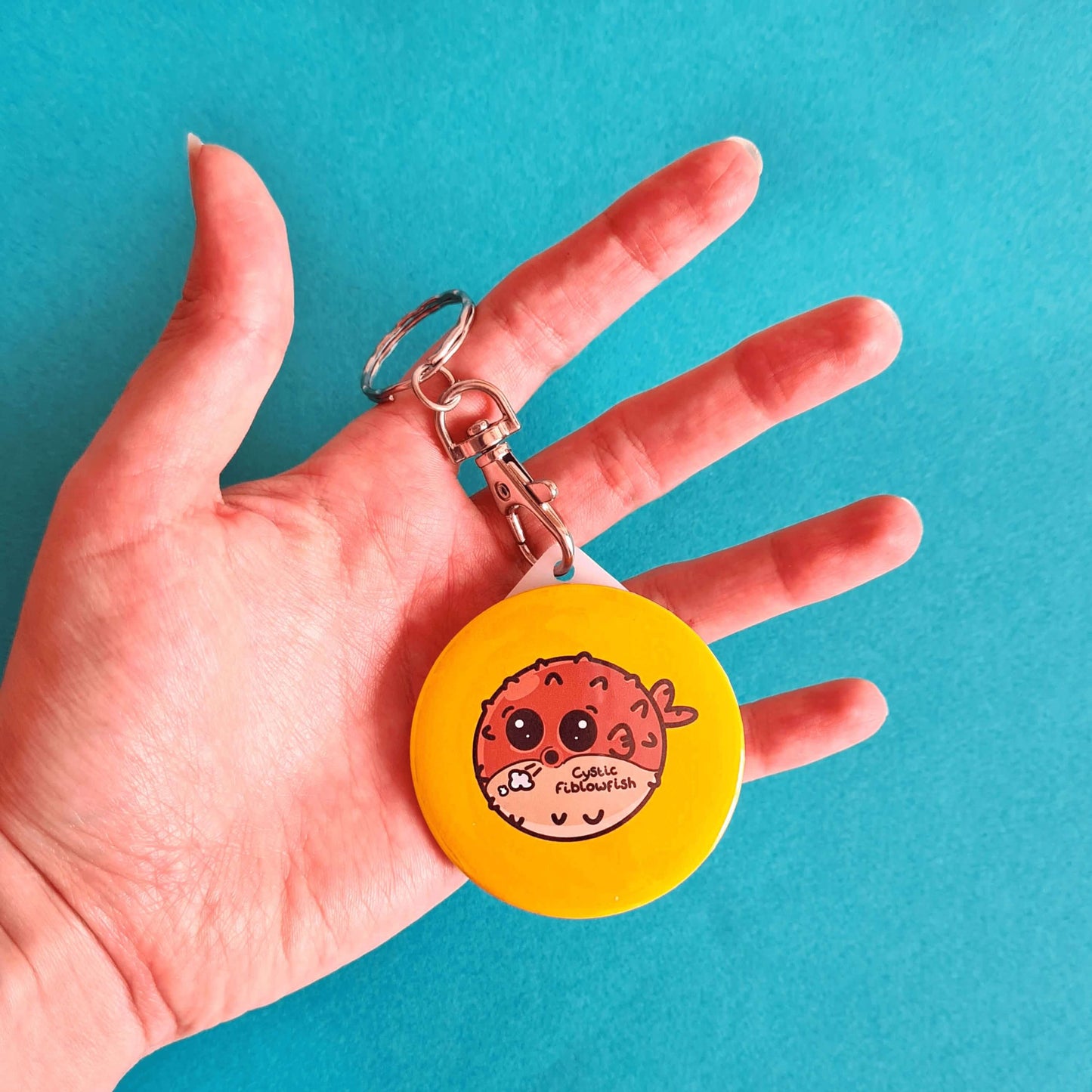 The Cystic Fiblowfish Keyring - Cystic Fibrosis being held on a blue background. The silver clip plastic yellow circle keychain with a puffer fish blowfish wheezing with 'cystic fiblowfish' written in black on its belly. The design is raising awareness for cystic fibrosis.