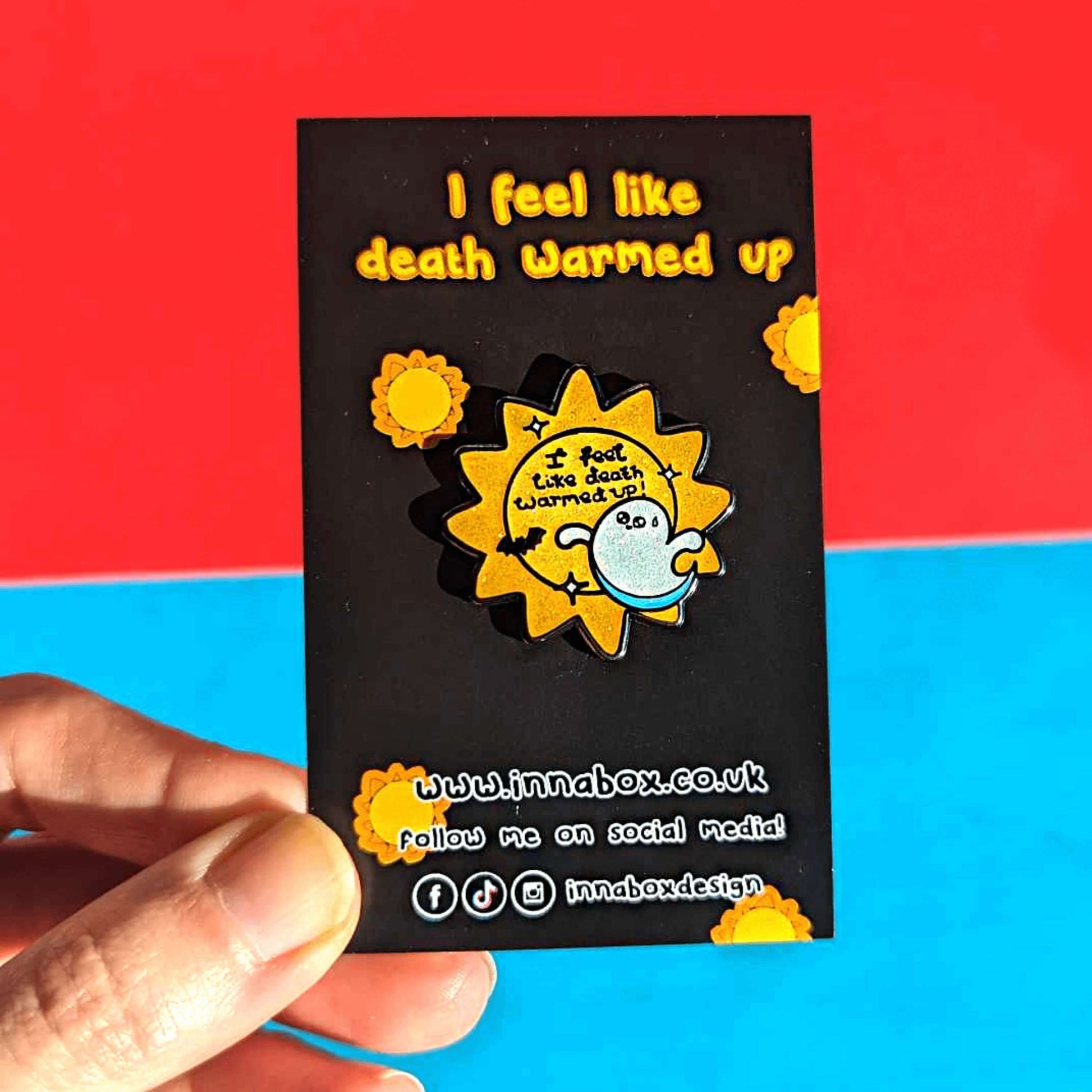 Hand holding Death Warmed Up Halloween Pin on black backing card with yellow sunshines, yellow top text reading 'I feel like death warmed up' and white bottom text of the innabox social media handles. The pin is glittery, yellow and sun shaped with 'I feel like death warmed up!' written in the middle of the sun in black writing and white sparkles around it. There is also a little black bat underneath the writing and a cute white ghost with big eyes, a little smile and a sweat droplet on it's forehead.