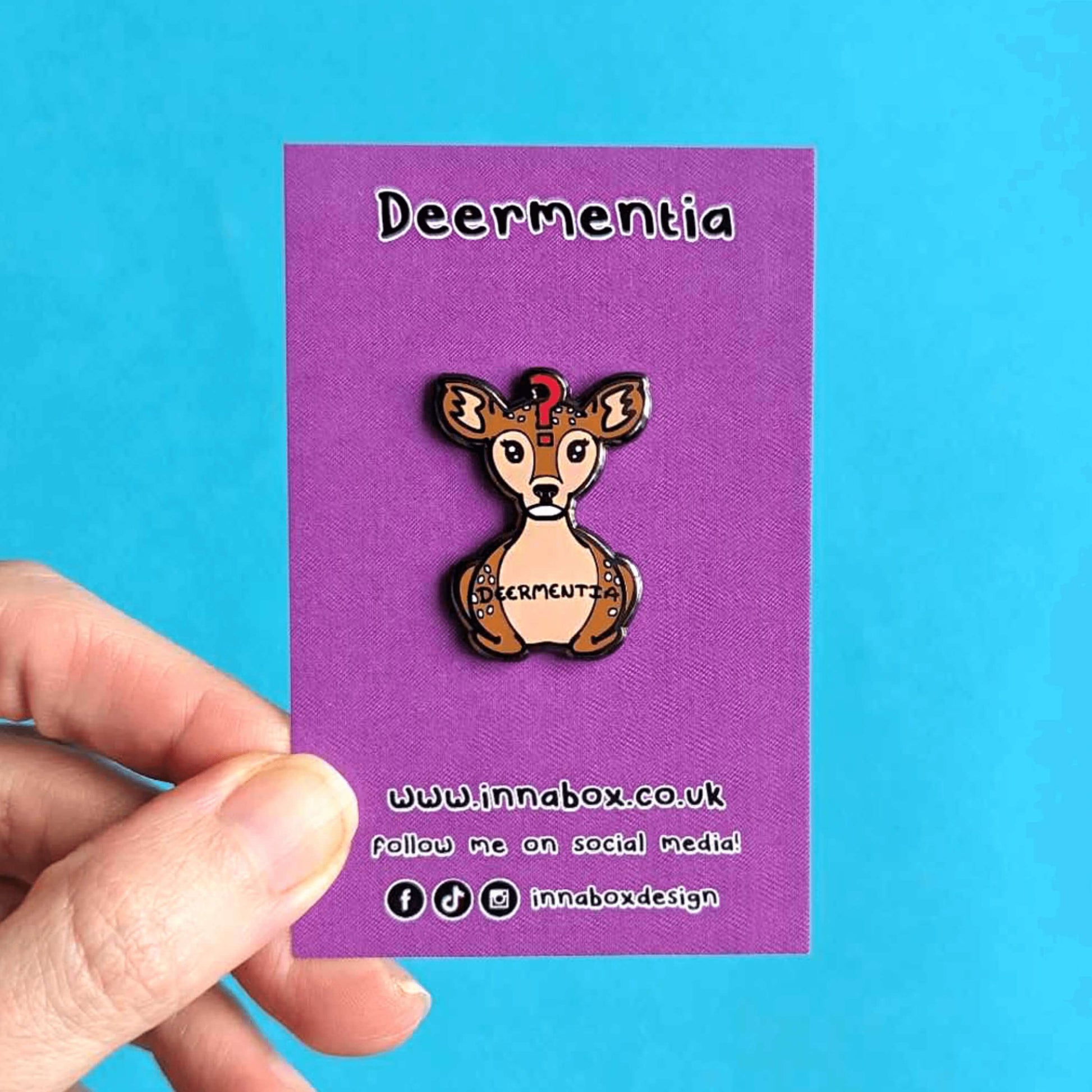 Hand holding Dementia enamel pin on it's purple backing in front of a blue background. A brown deer shaped enamel pin with long eyelashes, blank expression on it's face and red question mark in the middle of it's forehead. The deer has fluffy ears and white spots and is in a lying down straight on position. 'Deermentia' is written across the light brown tummy in black writing.