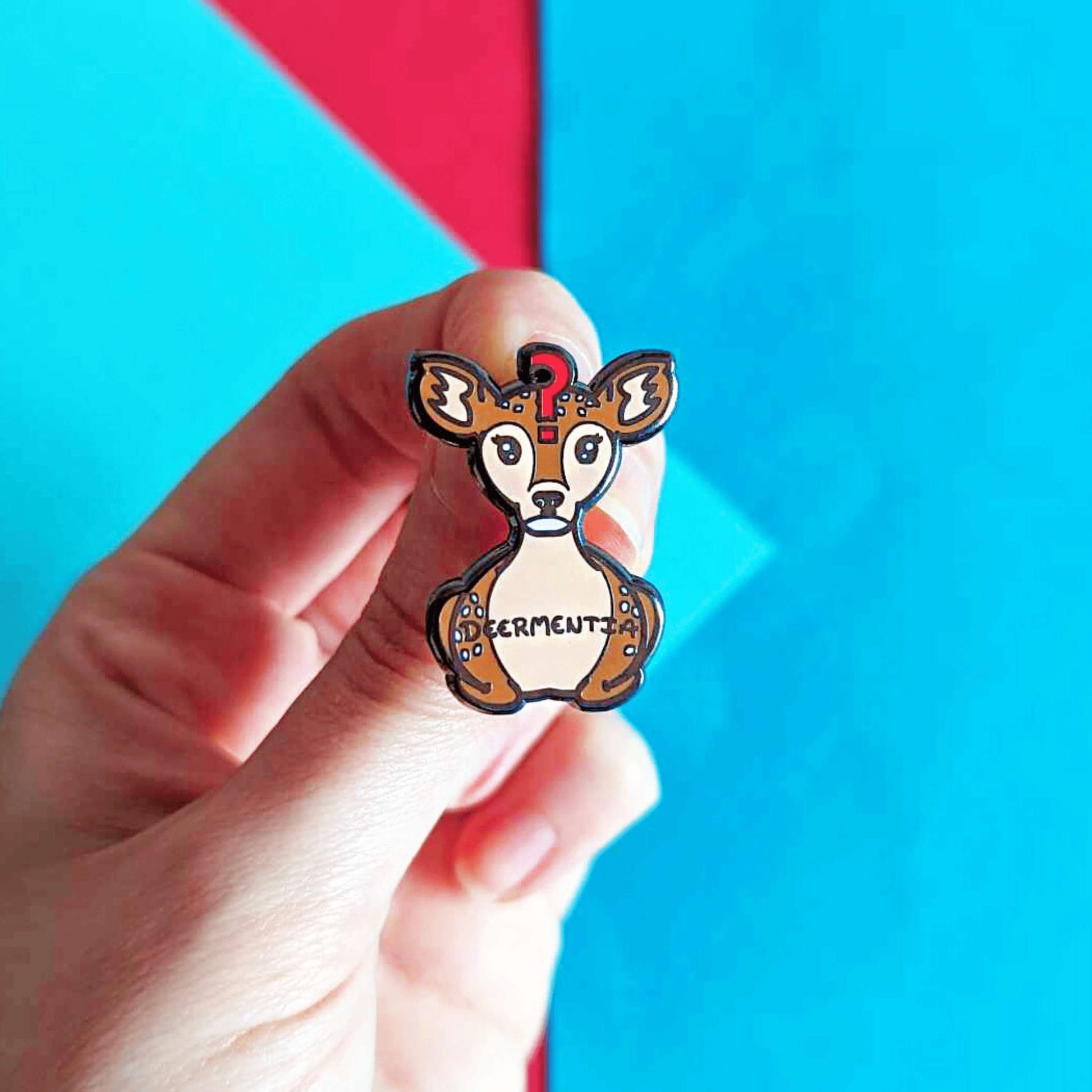 Hand holding Dementia enamel pin in front of red and blue background. A brown deer shaped enamel pin with long eyelashes, blank expression on it's face and red question mark in the middle of it's forehead. The deer has fluffy ears and white spots and is in a lying down straight on position. 'Deermentia' is written across the light brown tummy in black writing.