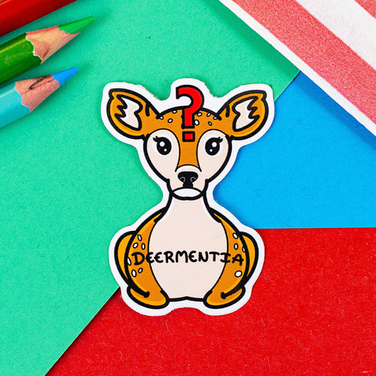 Deermentia Sticker - Dementia sticker shown on a red, blue and green background with colouring pencils and a red stripe candy bag. A brown deer shaped sticker with long eyelashes, blank expression on it's face and red question mark in the middle of it's forehead. The deer has fluffy ears and white spots and is in a lying down straight on position. 'Deermentia' is written across the light brown tummy in black writing. The design is to raise awareness for dementia.