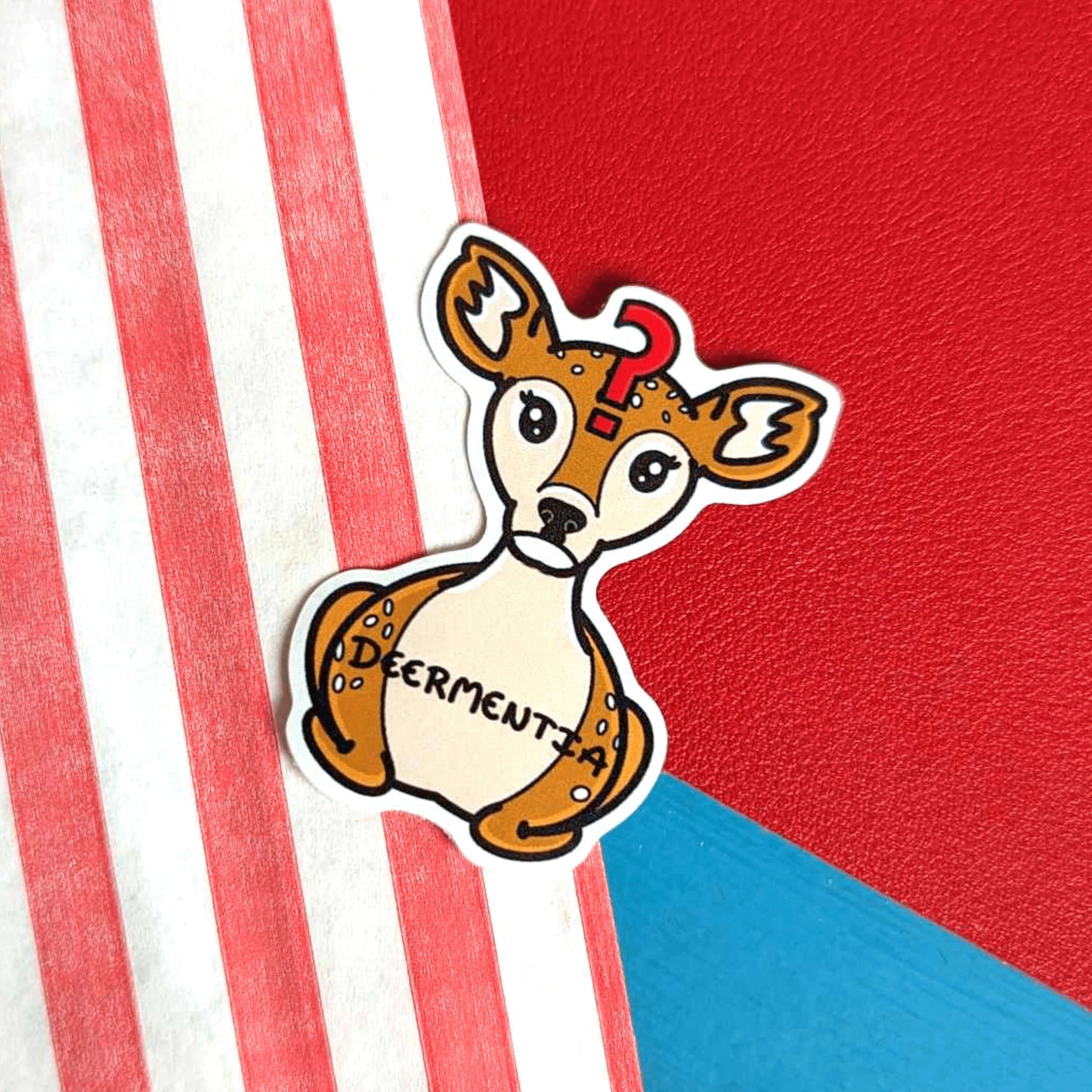 Deermentia Sticker - Dementia sticker shown on a red, white and blue background. A brown deer shaped sticker with long eyelashes, blank expression on it's face and red question mark in the middle of it's forehead. The deer has fluffy ears and white spots and is in a lying down straight on position. 'Deermentia' is written across the light brown tummy in black writing. The design is to raise awareness for dementia.
