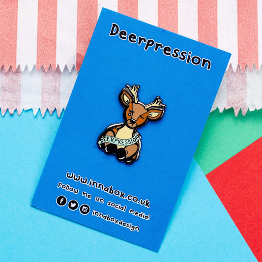The Deerpression Enamel Pin - Depression on blue backing card with black text above reading 'deerpression' and black bottom text of innabox social media handles, laid on a red, green and blue background. The deer shape pin its eyes closed with a blue tear falling from one eye, across its front is a blue banner with black text reading 'deerpression'. The design was created to raise awareness for depression.