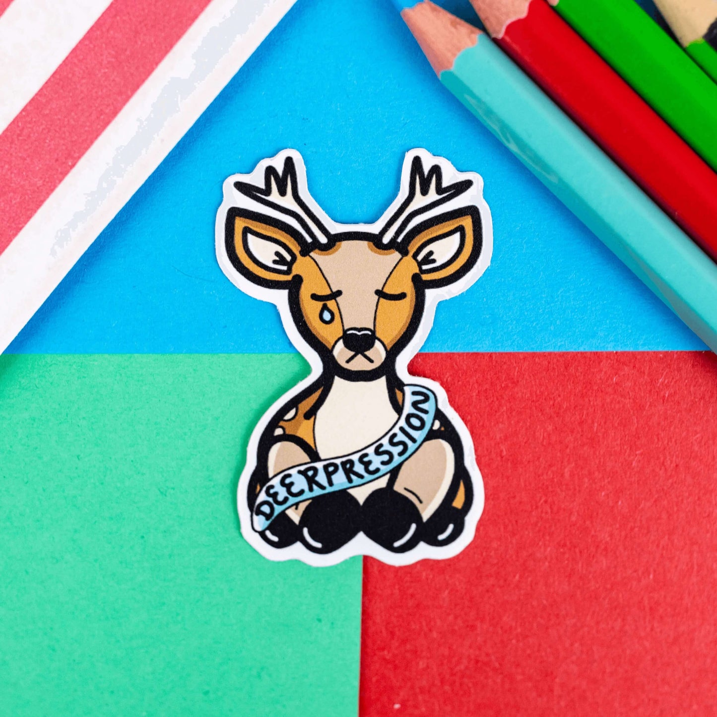 The Deerpression Sticker - Depression on a red, blue and green background with colouring pencils and a red stripe candy bag. The deer shaped sticker has its eyes closed with a tear falling down one cheek, across its middle is a blue banner with black text reading 'deerpression'. The design was created to raise awareness for depression.