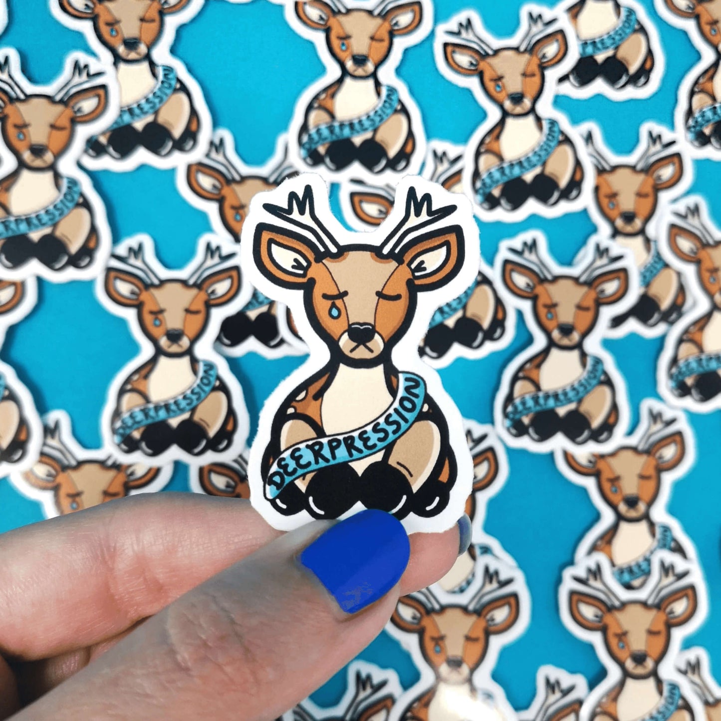 The Deerpression Sticker - Depression being held over multiple copies of the sticker on a blue background by a hand wearing blue nail varnish. The deer shaped sticker has its eyes closed with a tear falling down one cheek, across its middle is a blue banner with black text reading 'deerpression'. The design was created to raise awareness for depression.