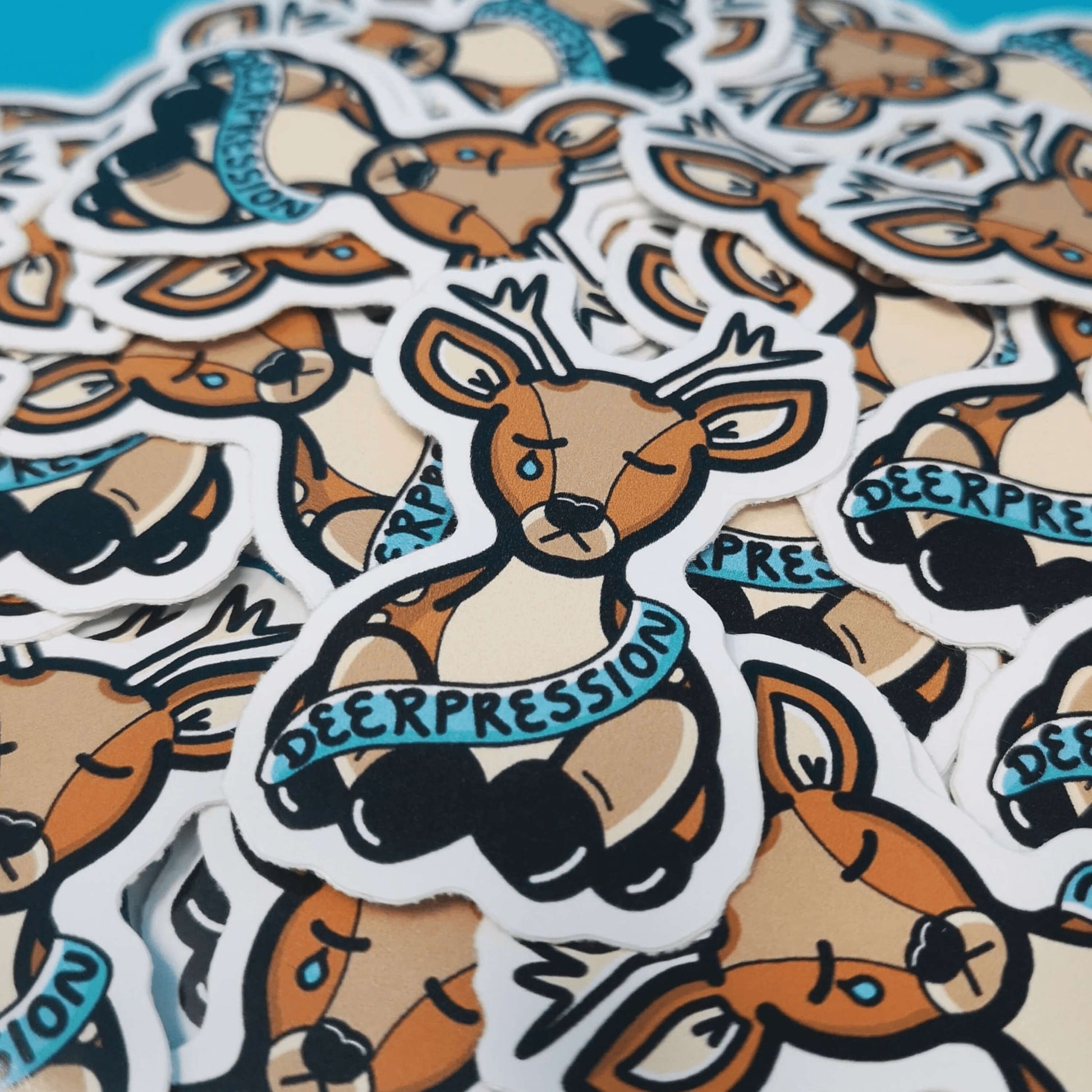 The Deerpression Sticker - Depression laying on a pile of multiple stickers on a blue background. The deer shaped sticker has its eyes closed with a tear falling down one cheek, across its middle is a blue banner with black text reading 'deerpression'. The design was created to raise awareness for depression.
