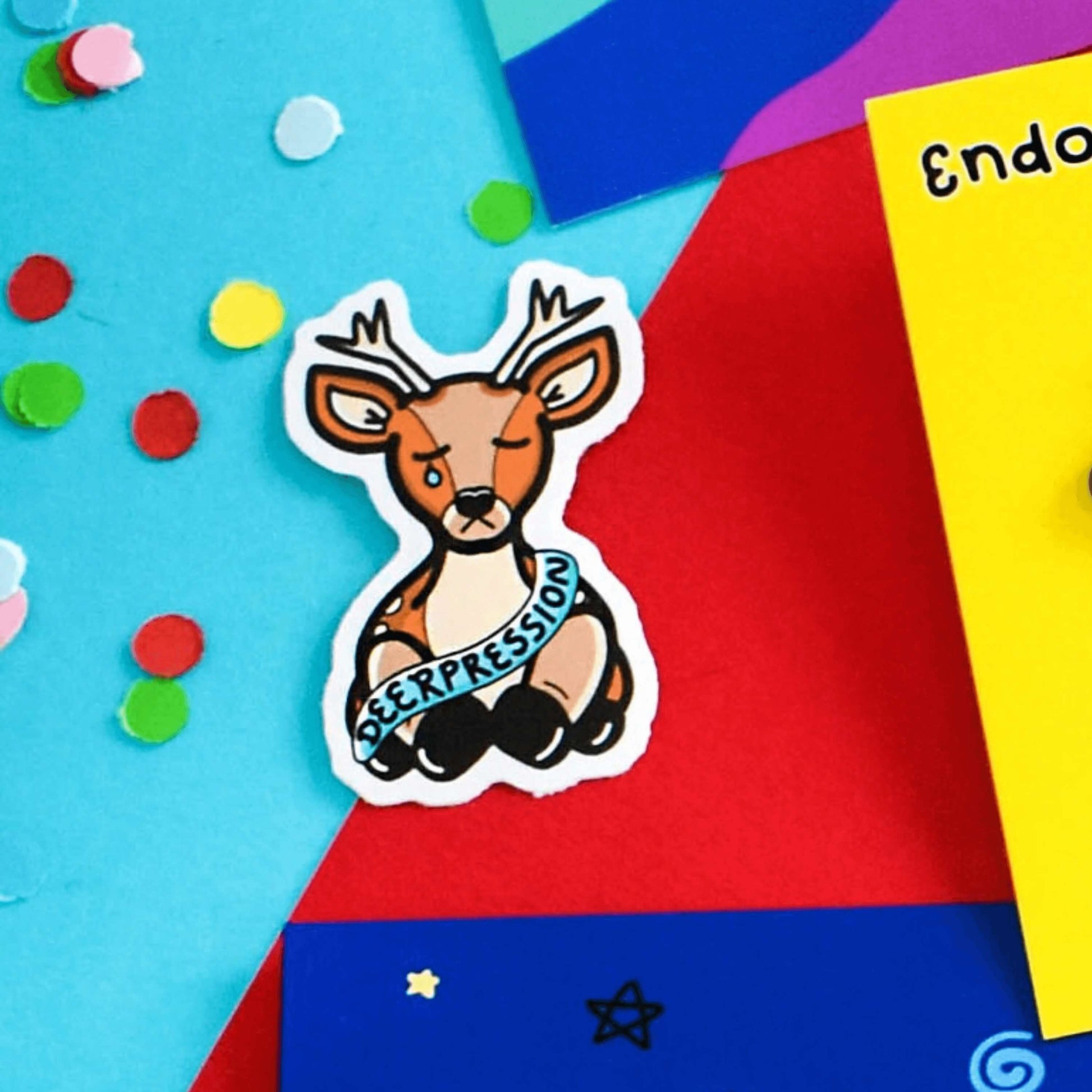 The Deerpression Sticker - Depression on a red and blue background with other innabox products and multicoloured confetti. The deer shaped sticker has its eyes closed with a tear falling down one cheek, across its middle is a blue banner with black text reading 'deerpression'. The design was created to raise awareness for depression.