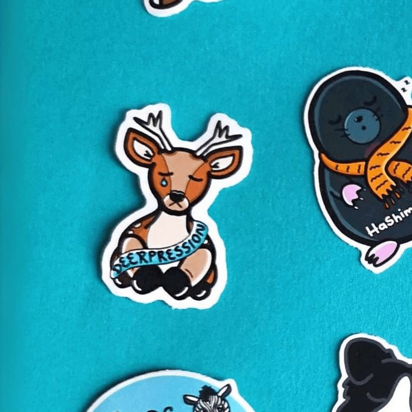 The Deerpression Sticker - Depression on a blue background with other innabox stickers. The deer shaped sticker has its eyes closed with a tear falling down one cheek, across its middle is a blue banner with black text reading 'deerpression'. The design was created to raise awareness for depression.