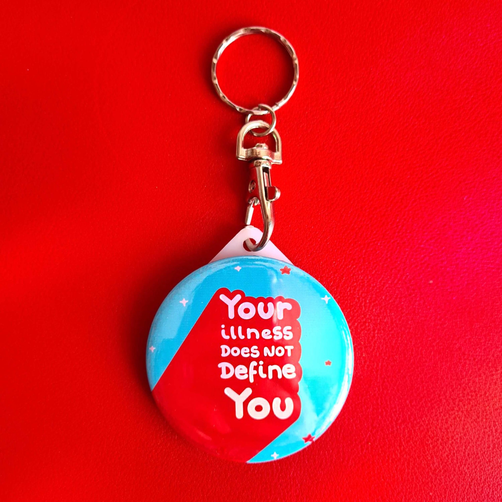 The Your Illness Does NOT Define You Keyring on a red background. The silver clip blue plastic circular keyring has red and white sparkles and text reading 'your illness does not define you'. A hand drawn design raising awareness for invisible illnesses.