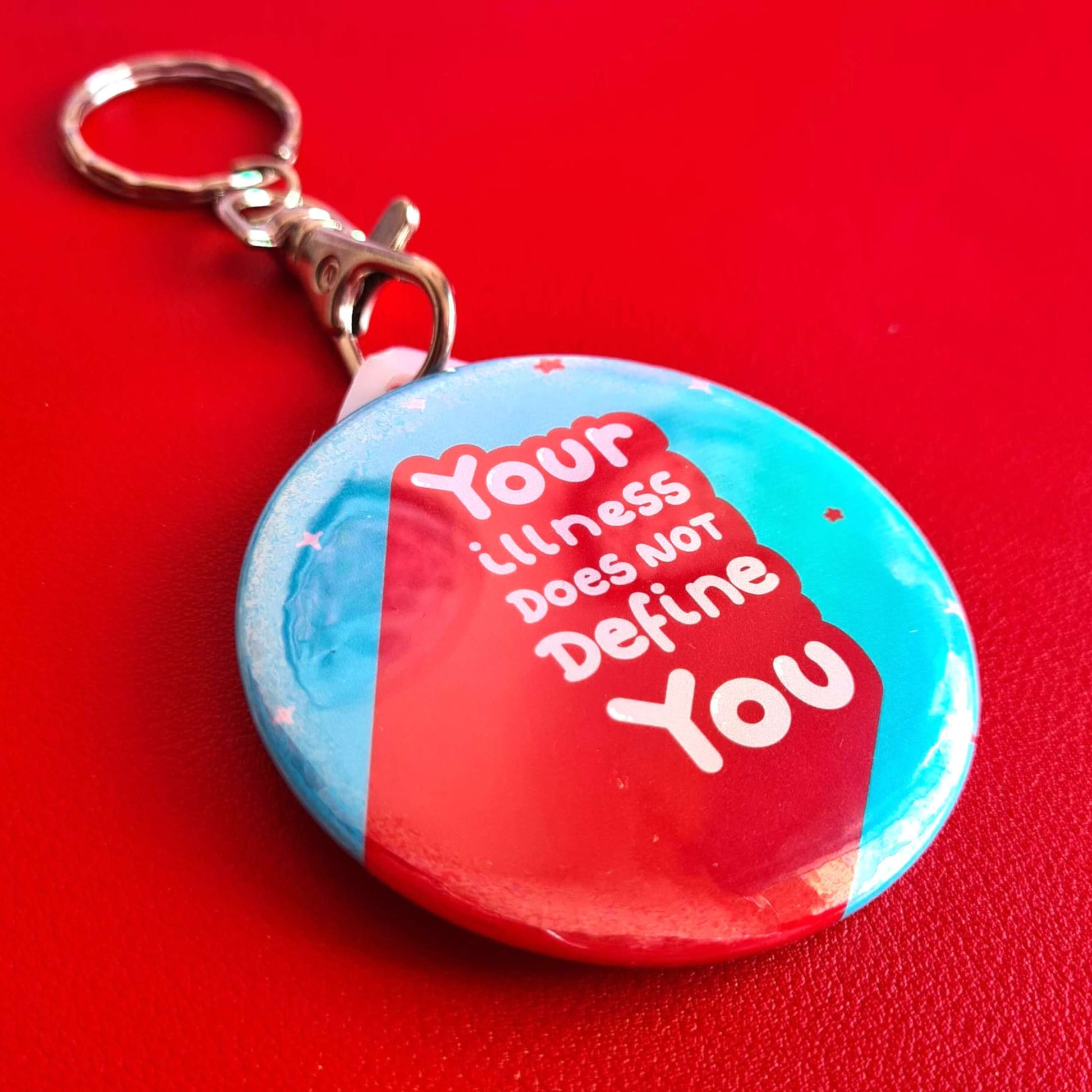 The Your Illness Does NOT Define You Keyring on a red background. The silver clip blue plastic circular keyring has red and white sparkles and text reading 'your illness does not define you'. A hand drawn design raising awareness for invisible illnesses.