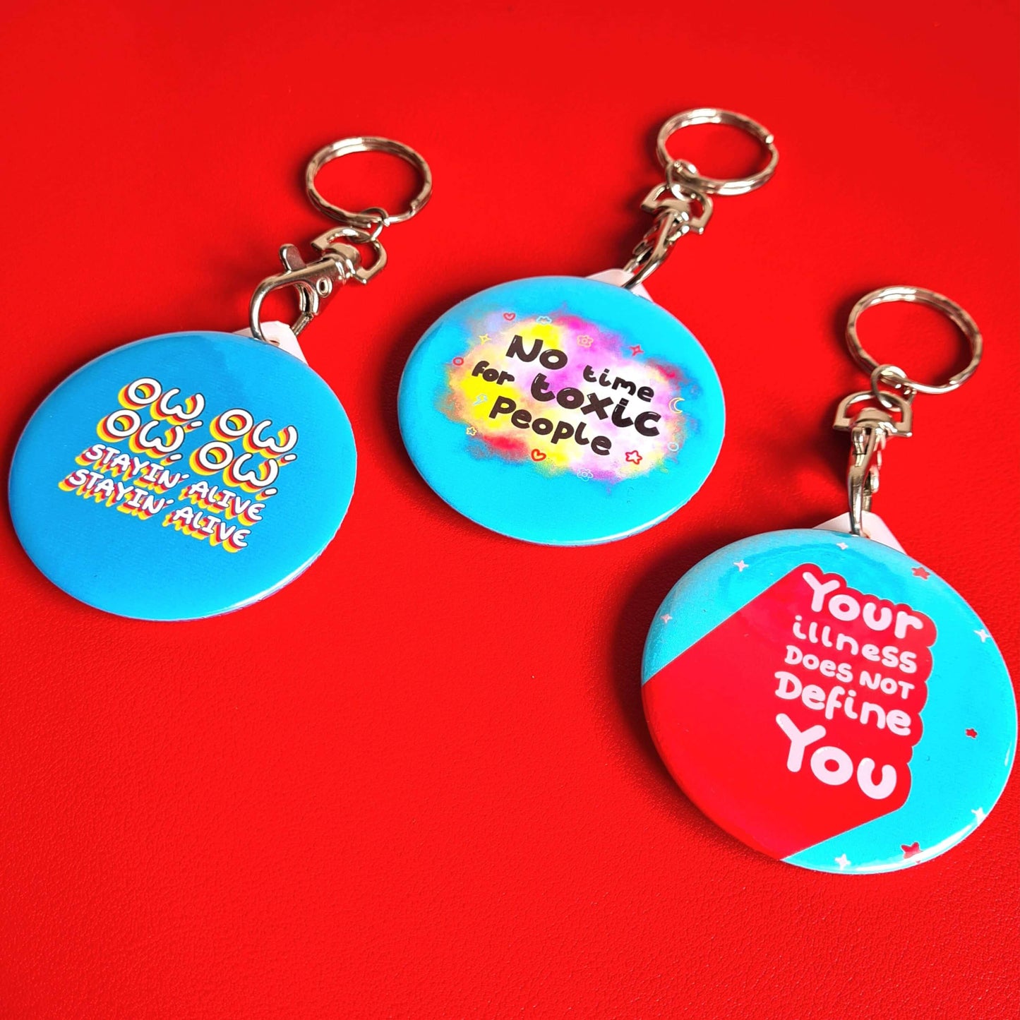 The Your Illness Does NOT Define You Keyring on a red background with other innabox keychains. The silver clip blue plastic circular keyring has red and white sparkles and text reading 'your illness does not define you'. A hand drawn design raising awareness for invisible illnesses.