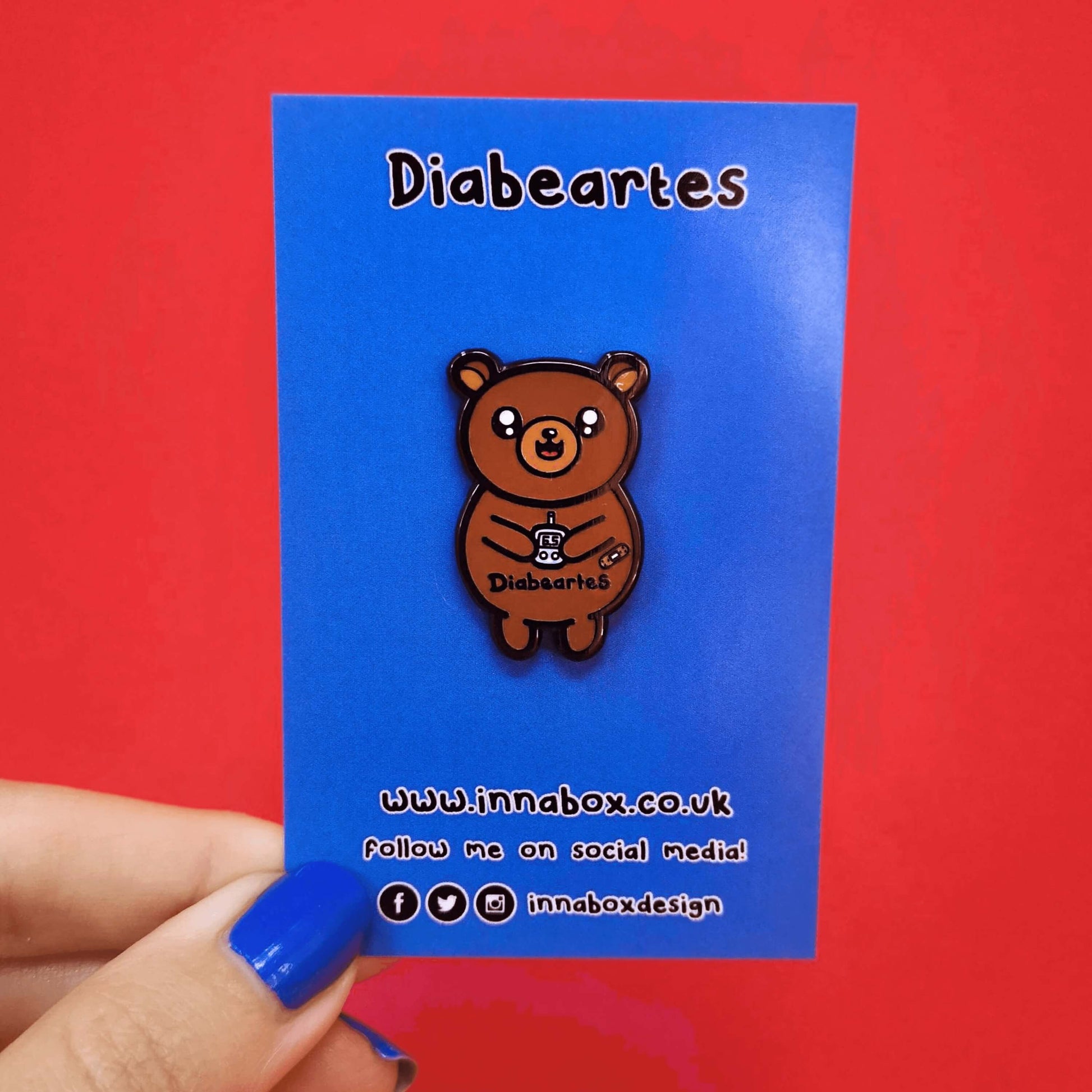 The Diabeartes Enamel Pin - Diabetes on blue backing card with black text above reading 'diabeartes' and below text of the innabox website and social handles being held over a red background. The brown bear shaped pin is smiling holding a blood glucose reader with a plaster on its arm, across its tummy reads 'diabeartes' in black. The design is raising awareness for those with type 1 diabetes and type 2 diabetes.