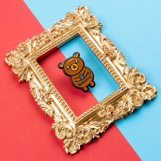  The Diabeartes Enamel Pin - Diabetes on red and blue card inside a gold ornate frame. The brown bear shaped pin is smiling holding a blood glucose reader with a plaster on its arm, across its tummy reads 'diabeartes' in black. The design is raising awareness for those with type 1 diabetes and type 2 diabetes.