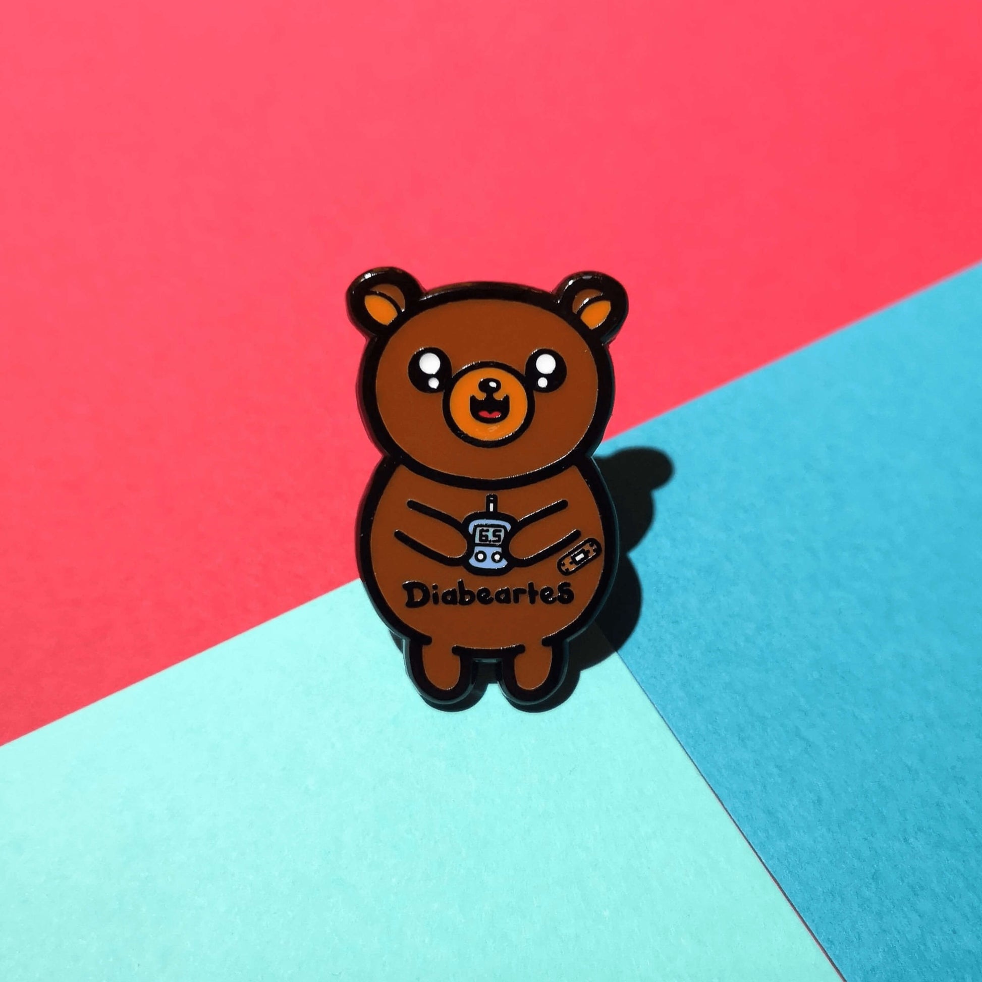 The Diabeartes Enamel Pin - Diabetes on a red and blue background. The brown bear shaped pin is smiling holding a blood glucose reader with a plaster on its arm, across its tummy reads 'diabeartes' in black. The design is raising awareness for those with type 1 diabetes and type 2 diabetes.