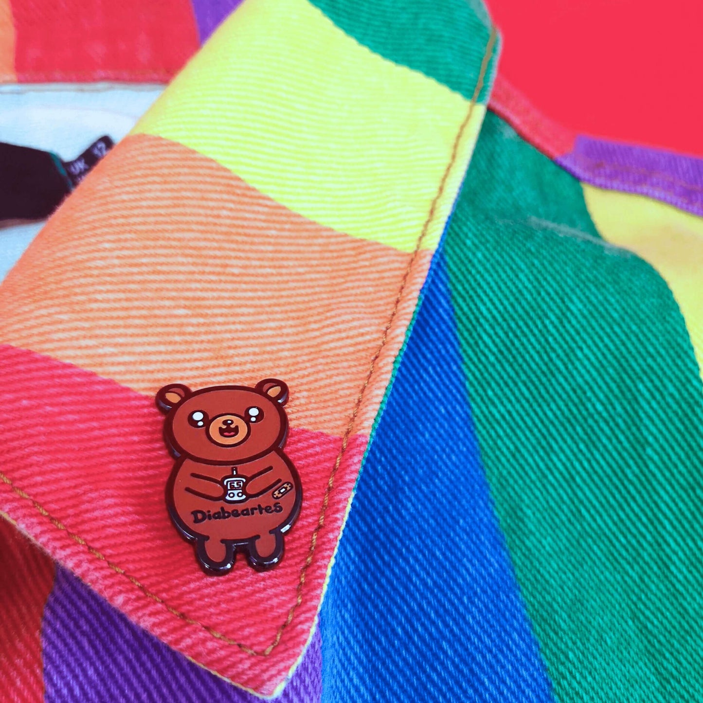 The Diabeartes Enamel Pin - Diabetes pinned onto the collar of a rainbow denim jacket. The brown bear shaped pin is smiling holding a blood glucose reader with a plaster on its arm, across its tummy reads 'diabeartes' in black. The design is raising awareness for those with type 1 diabetes and type 2 diabetes.