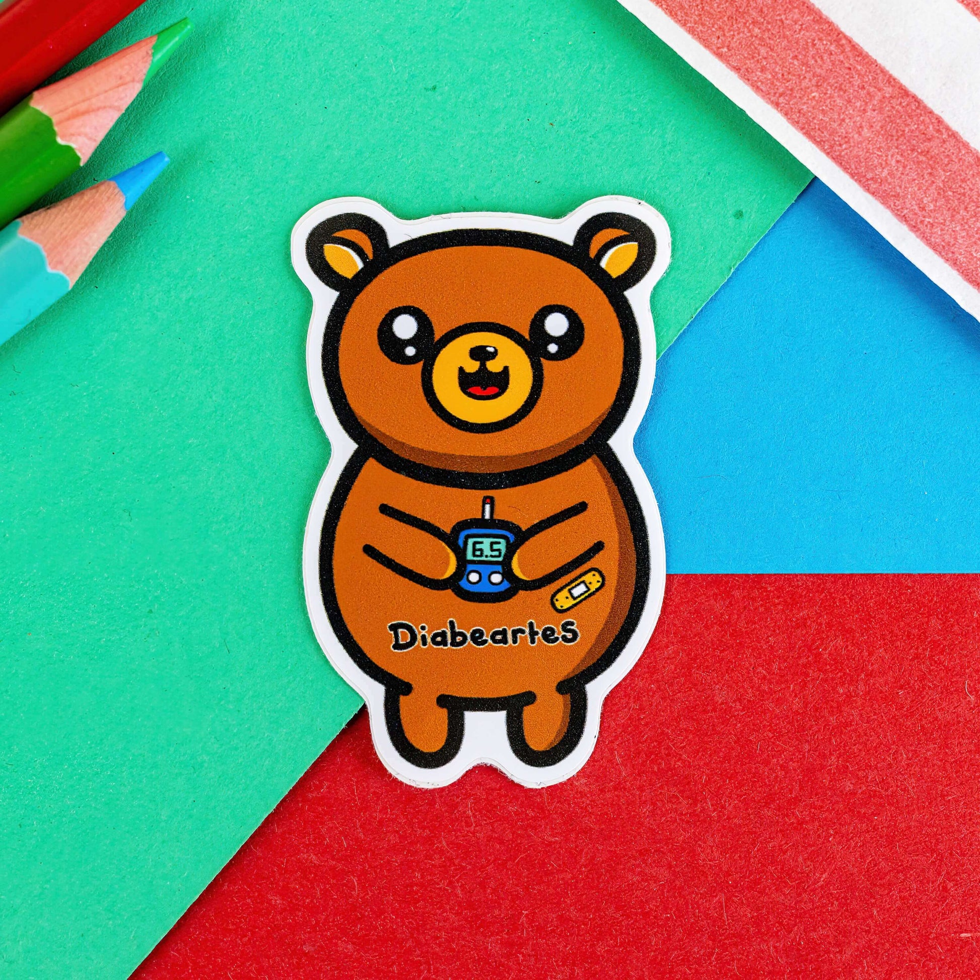 The Diabeartes Sticker - Diabetes on a red, green and blue background with colouring pencils and a red stripe candy bag. The brown bear shaped sticker is smiling holding a blood glucose reader with a plaster on its arm, across its tummy reads 'diabeartes' in black. The design is raising awareness for those with type 1 diabetes and type 2 diabetes.