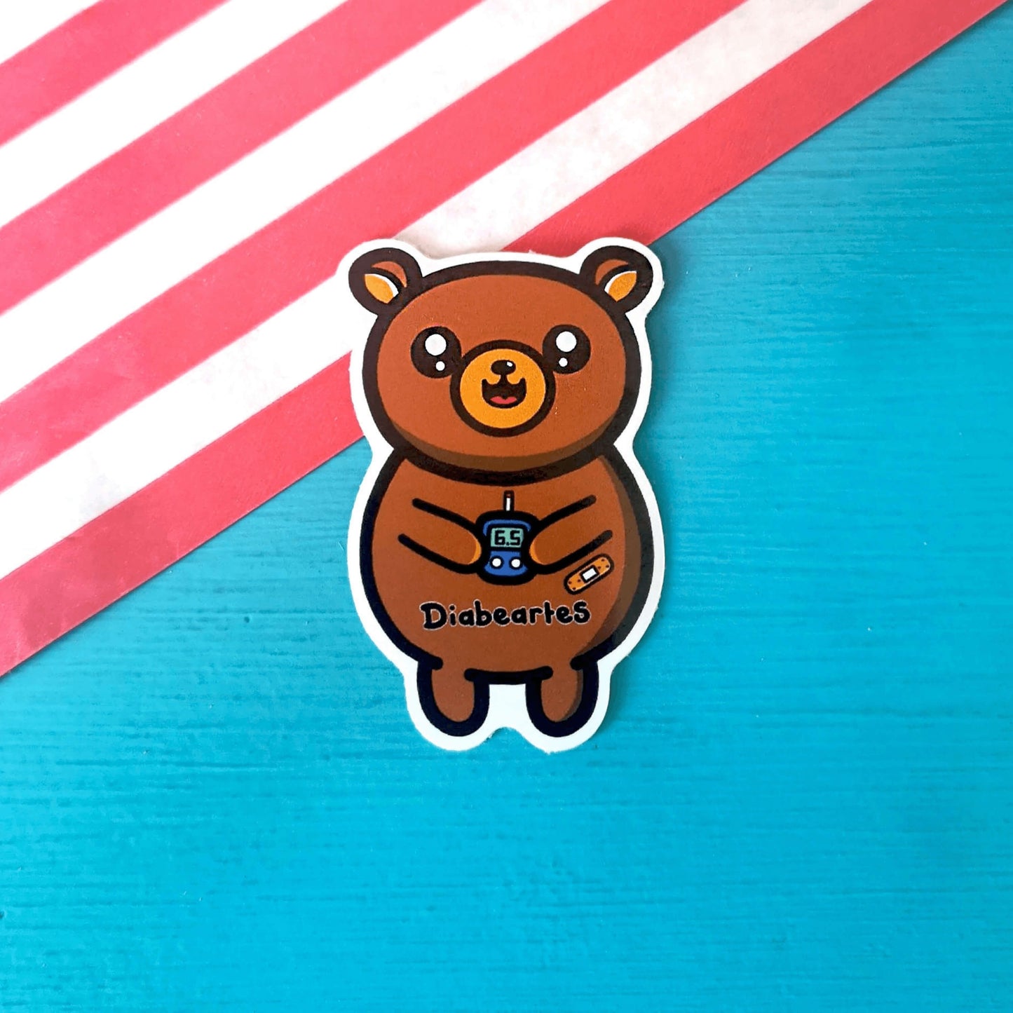 The Diabeartes Sticker - Diabetes on a red stripe candy bag and blue background. The brown bear shaped sticker is smiling holding a blood glucose reader with a plaster on its arm, across its tummy reads 'diabeartes' in black. The design is raising awareness for those with type 1 diabetes and type 2 diabetes.