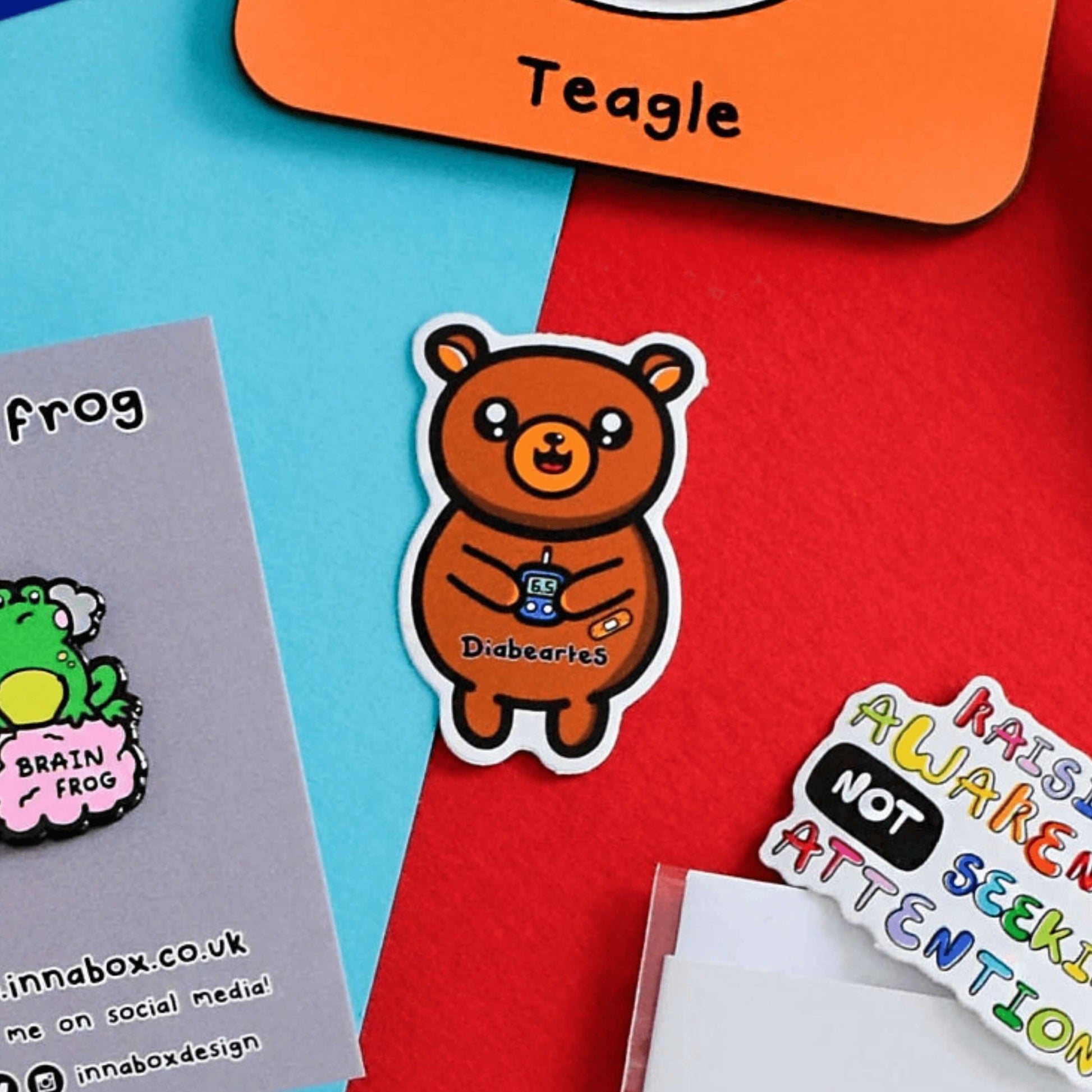 The Diabeartes Sticker - Diabetes on a red and blue background with other innabox products. The brown bear shaped sticker is smiling holding a blood glucose reader with a plaster on its arm, across its tummy reads 'diabeartes' in black. The design is raising awareness for those with type 1 diabetes and type 2 diabetes.