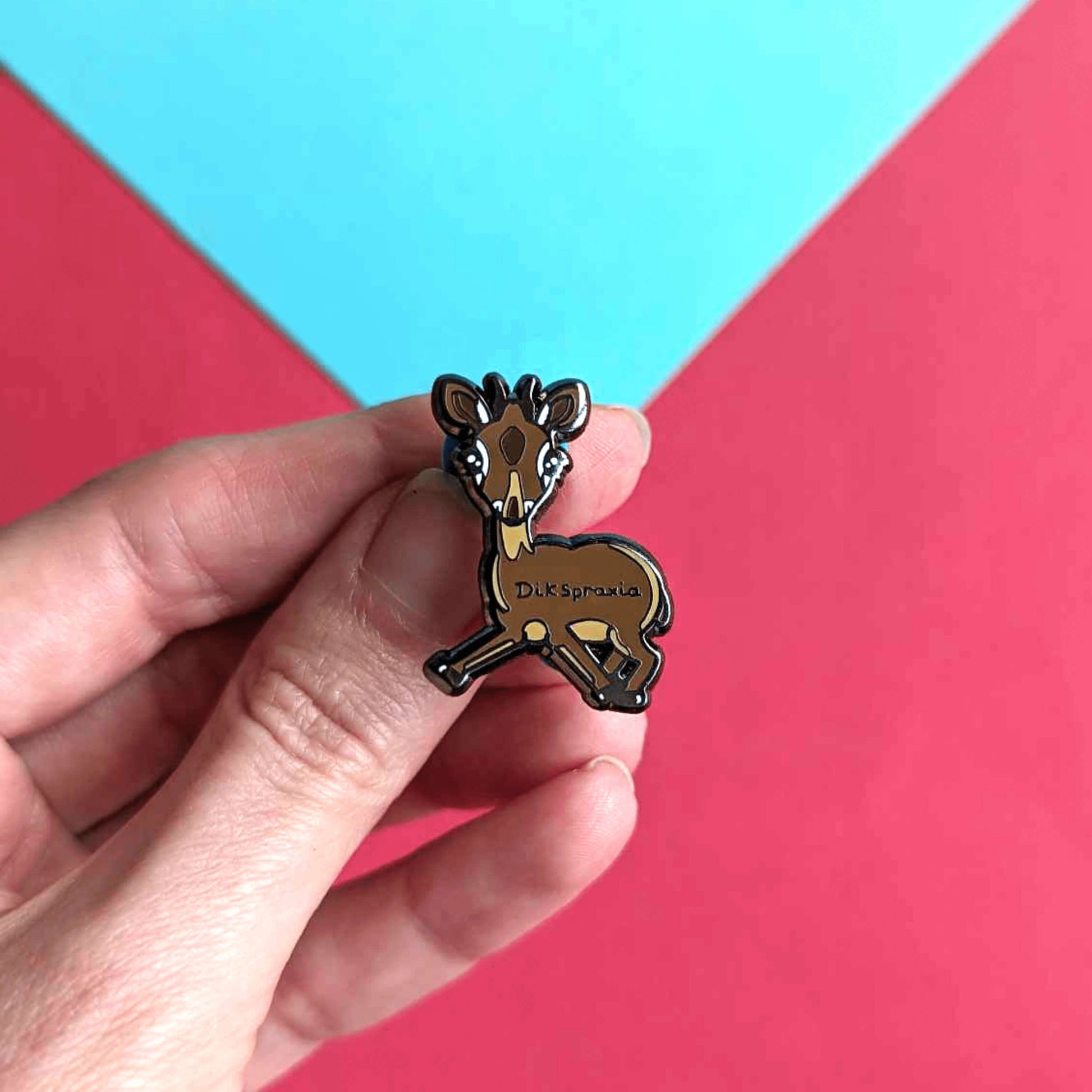 The Dikspraxia Enamel Pin - Dyspraxia being held up over a red and blue background. The brown dik dik antelope shaped enamel pin has its two front legs splayed chaotically with black text reading 'dikspraxia' across its middle. The design is raising awareness for dyspraxia and neurodivergence.
