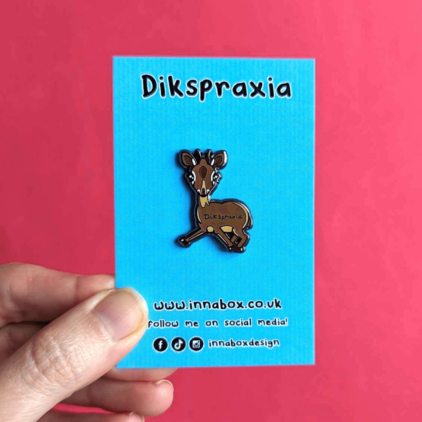 The Dikspraxia Enamel Pin - Dyspraxia on blue backing card with top text reading 'dikspraxia' and bottom text of the innabox website and social media handles being held up over a red background. The brown dik dik antelope shaped enamel pin has its two front legs splayed chaotically with black text reading 'dikspraxia' across its middle. The design is raising awareness for dyspraxia and neurodivergence.