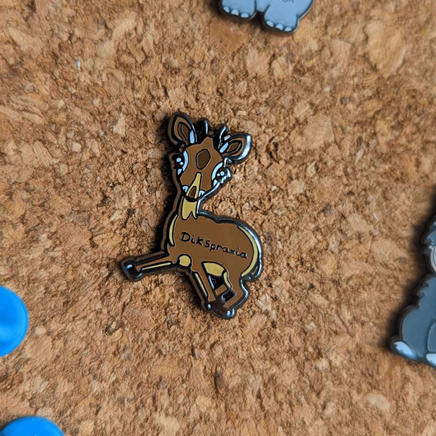 The Dikspraxia Enamel Pin - Dyspraxia pinned on a brown corkboard with other innabox pins. The brown dik dik antelope shaped enamel pin has its two front legs splayed chaotically with black text reading 'dikspraxia' across its middle. The design is raising awareness for dyspraxia and neurodivergence.