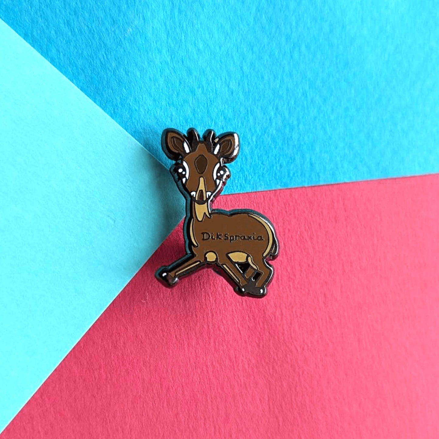 The Dikspraxia Enamel Pin - Dyspraxia on a red and blue background. The brown dik dik antelope shaped enamel pin has its two front legs splayed chaotically with black text reading 'dikspraxia' across its middle. The design is raising awareness for dyspraxia and neurodivergence.