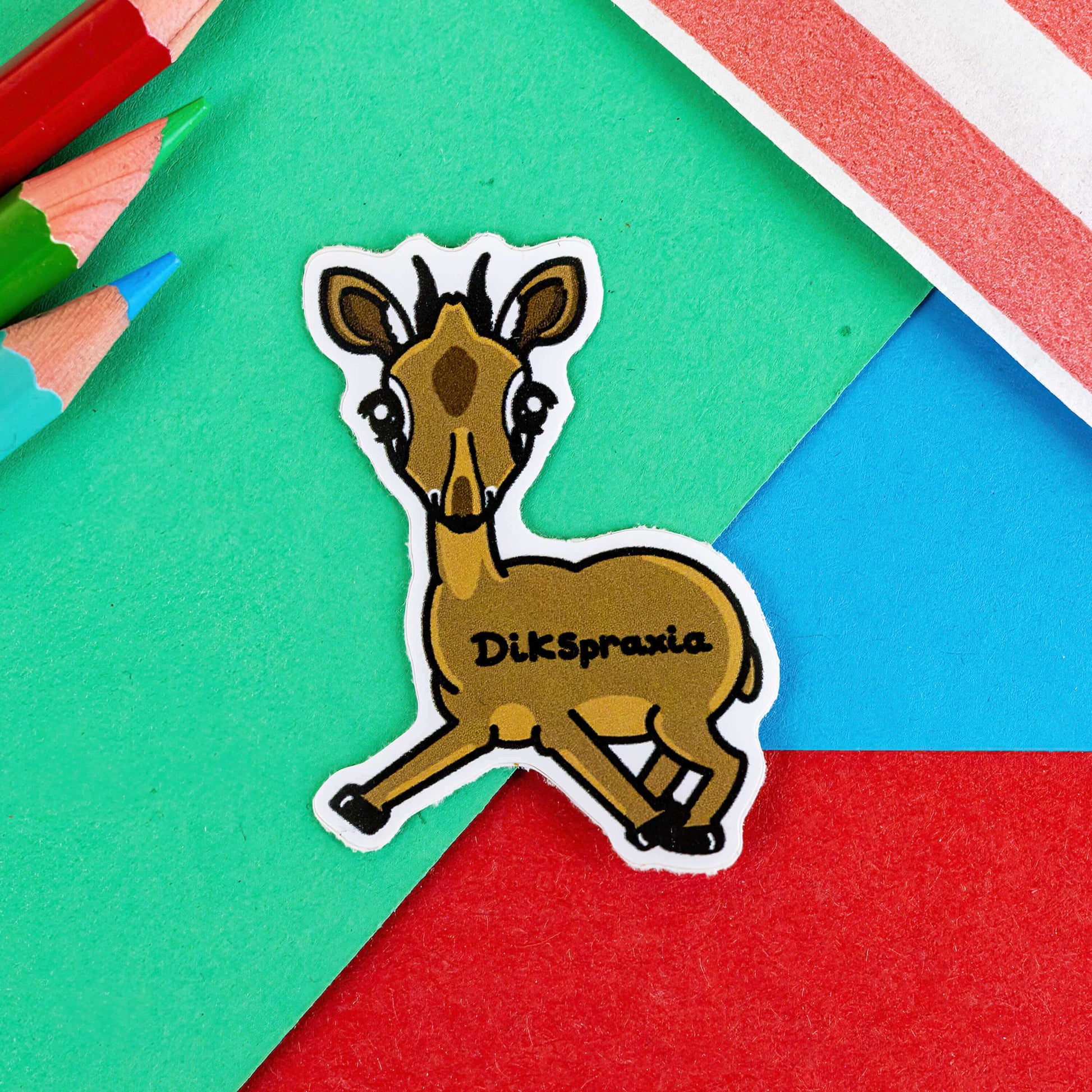 The Dikspraxia Sticker - Dyspraxia on a red, blue and green background with a red stripe candy bag and colouring pencils. The brown dik dik antelope shaped sticker has its two front legs splayed chaotically with black text reading 'dikspraxia' across its middle. The design is raising awareness for dyspraxia and neurodivergence.