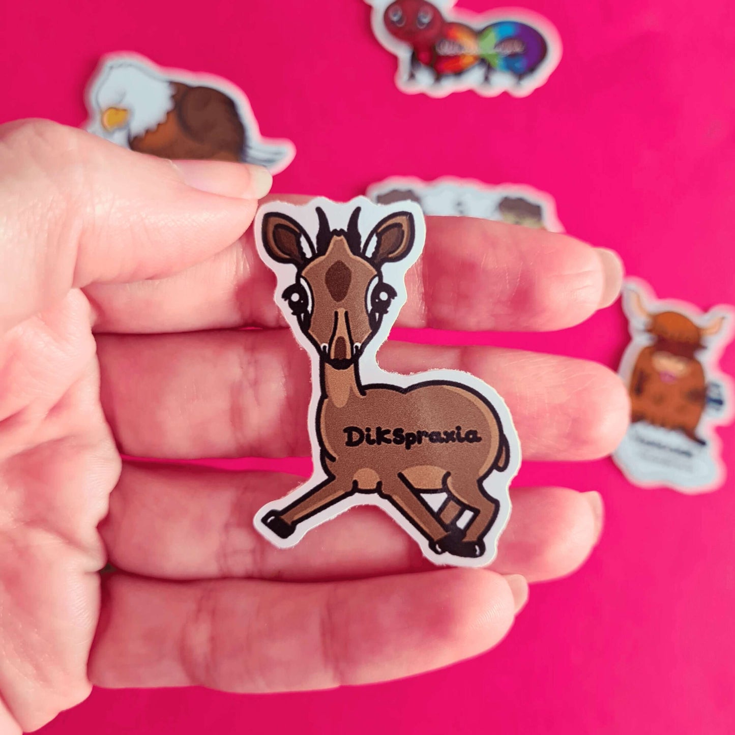 The Dikspraxia Sticker - Dyspraxia held over a red background with other innabox stickers. The brown dik dik antelope shaped sticker has its two front legs splayed chaotically with black text reading 'dikspraxia' across its middle. The design is raising awareness for dyspraxia and neurodivergence.
