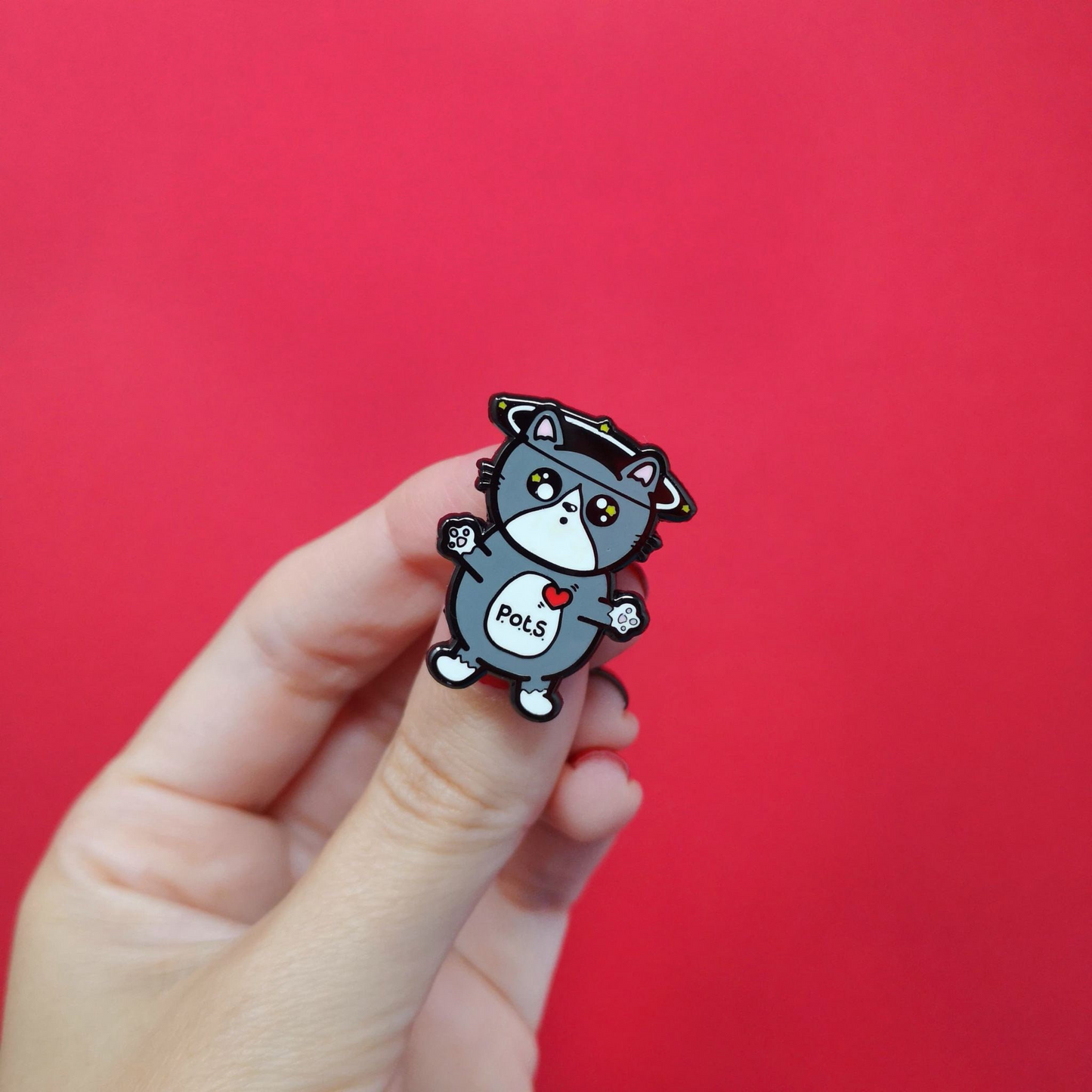 An enamel pin of a cat illustration with spinning dizzy stars around its head with its paws outstretched and postural tachycatdid syndrome (pots) written on its belly it is on a red background with a hand holding it. Postural tachycardia syndrome (PoTS) is when your heart rate increases very quickly after getting up from sitting or lying down.