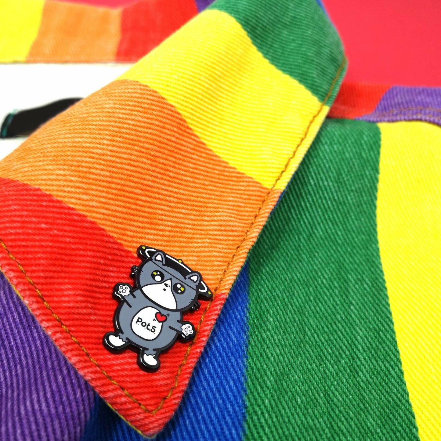 An enamel pin of a cat illustration with spinning dizzy stars around its head with its paws outstretched and postural tachycatdid syndrome (pots) written on its belly it is on a rainbow denim jacket collar. Postural tachycardia syndrome (PoTS) is when your heart rate increases very quickly after getting up from sitting or lying down.
