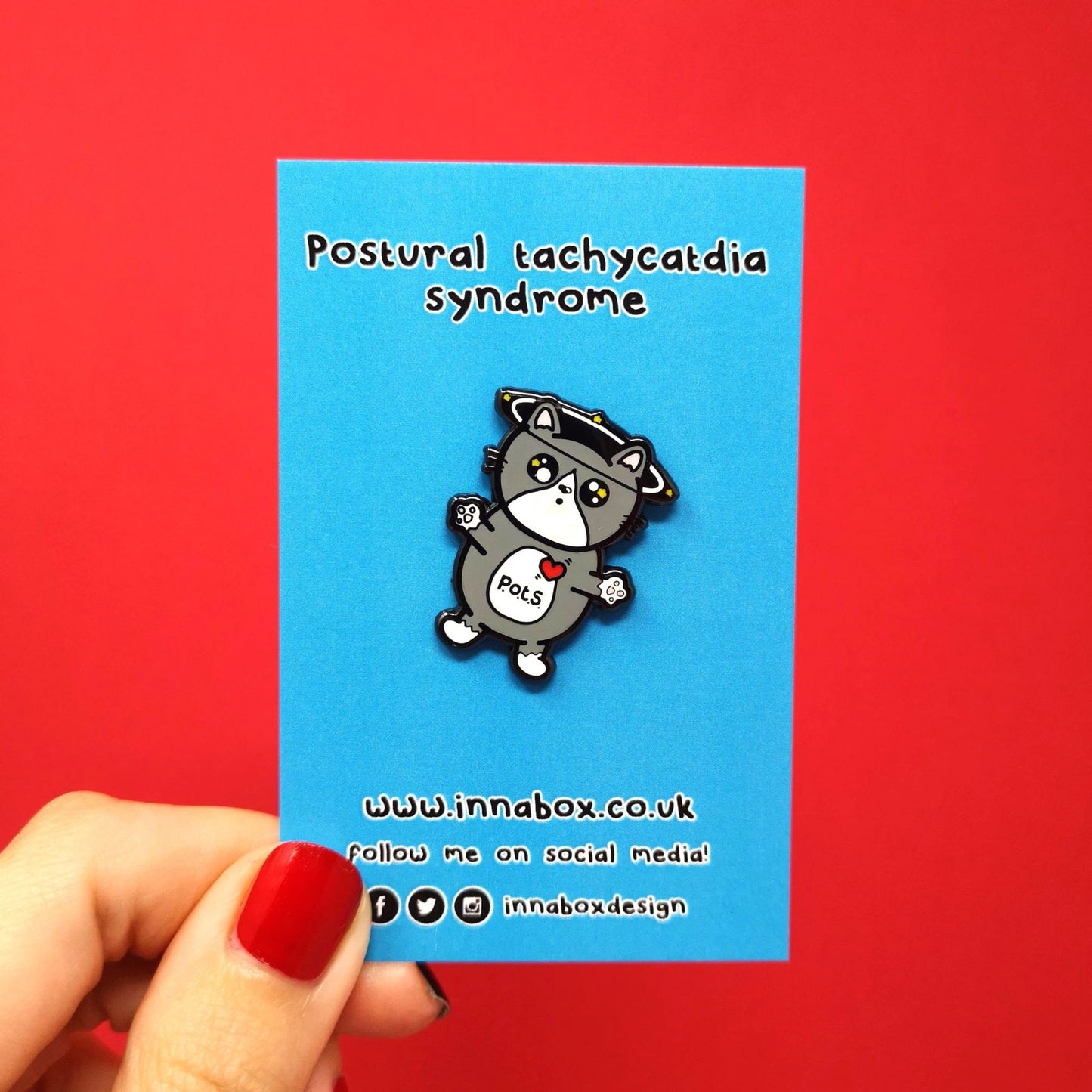 An enamel pin of a cat illustration with spinning dizzy stars around its head with its paws outstretched and postural tachycatdid syndrome (pots) written on its belly it is on a red background on a blue backing card with a hand holding it. Postural tachycardia syndrome (PoTS) is when your heart rate increases very quickly after getting up from sitting or lying down.