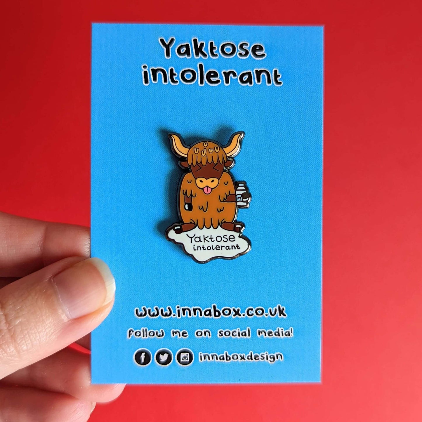Yaktose Intolerant Enamel Pin - Lactose Intolerant on blue backing card held in front of a red background. The enamel pin is a yak sat holding a bottle of milk with it's tongue out looking disgusted. There is a puddle of milk under the yak with black text that reads 'yaktose intolerant'. The hand drawn design is made to raise awareness for lactose intolerance