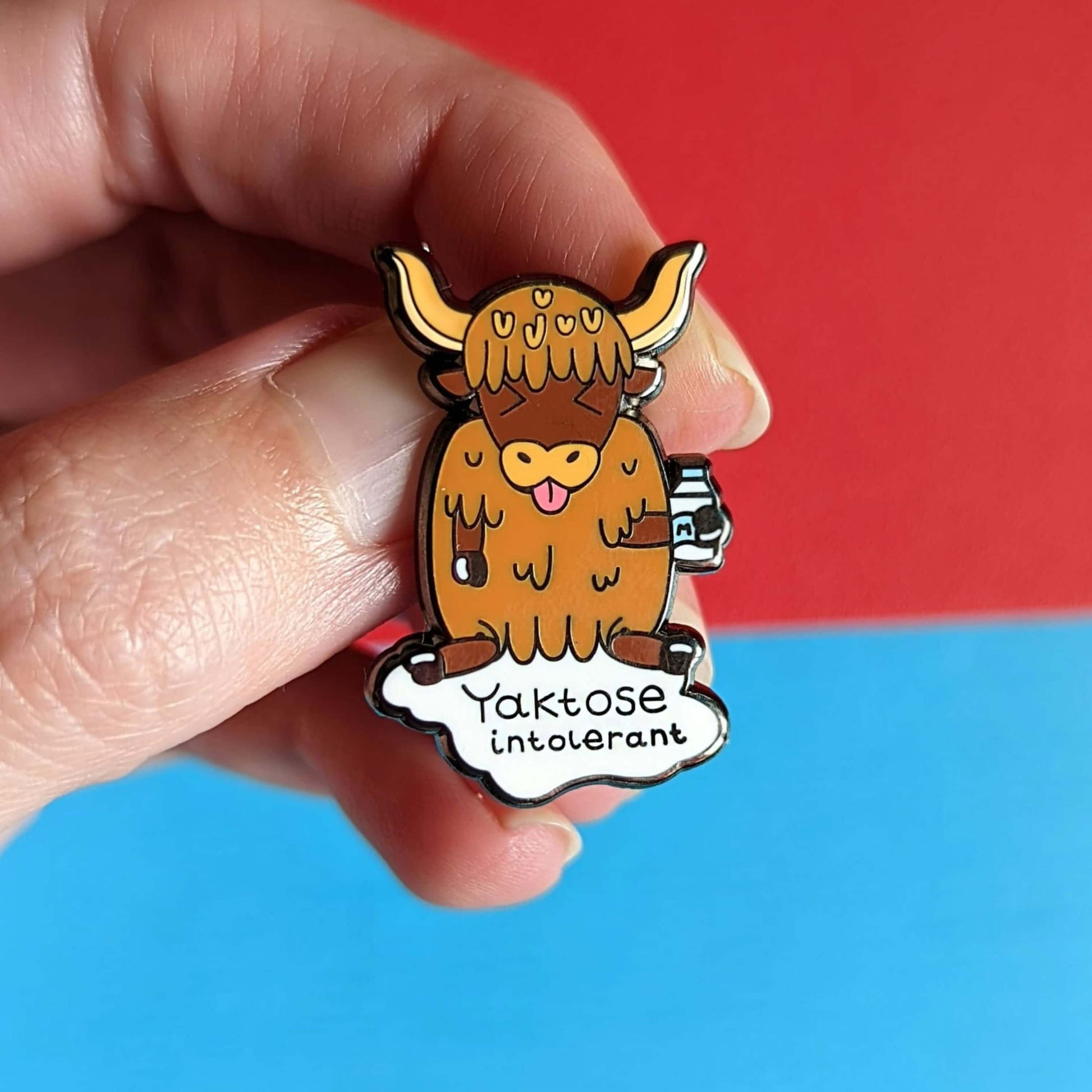 Yaktose Intolerant Enamel Pin - Lactose Intolerant held in front of a blue and red background. The enamel pin is a yak sat holding a bottle of milk with it's tongue out looking disgusted. There is a puddle of milk under the yak with black text that reads 'yaktose intolerant'. The hand drawn design is made to raise awareness for lactose intolerance